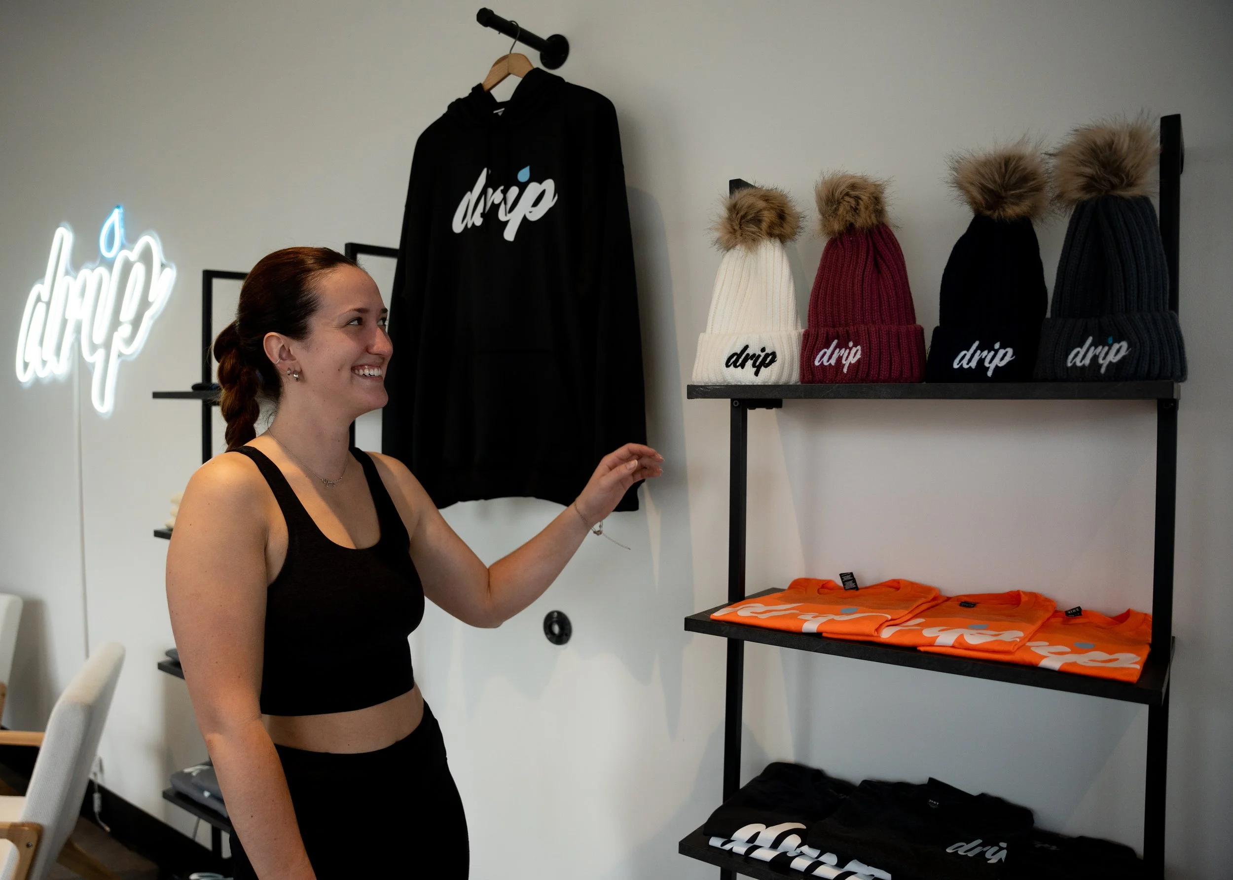 A woman with a ponytail and sleeveless black top smiling and reaching out to a display of clothing and hats inside a store. The store has a white wall, a neon sign with the word 'drip' and a black and white logo, and shelves with beanies and T-shirts.