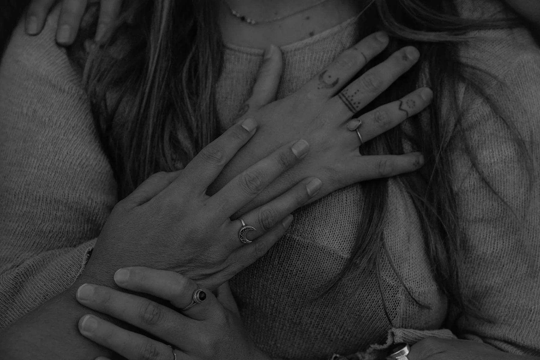 Close-up of two hands with rings, one on top of the other, placed on a person's chest; tattoos on fingers and rings on fingers.
