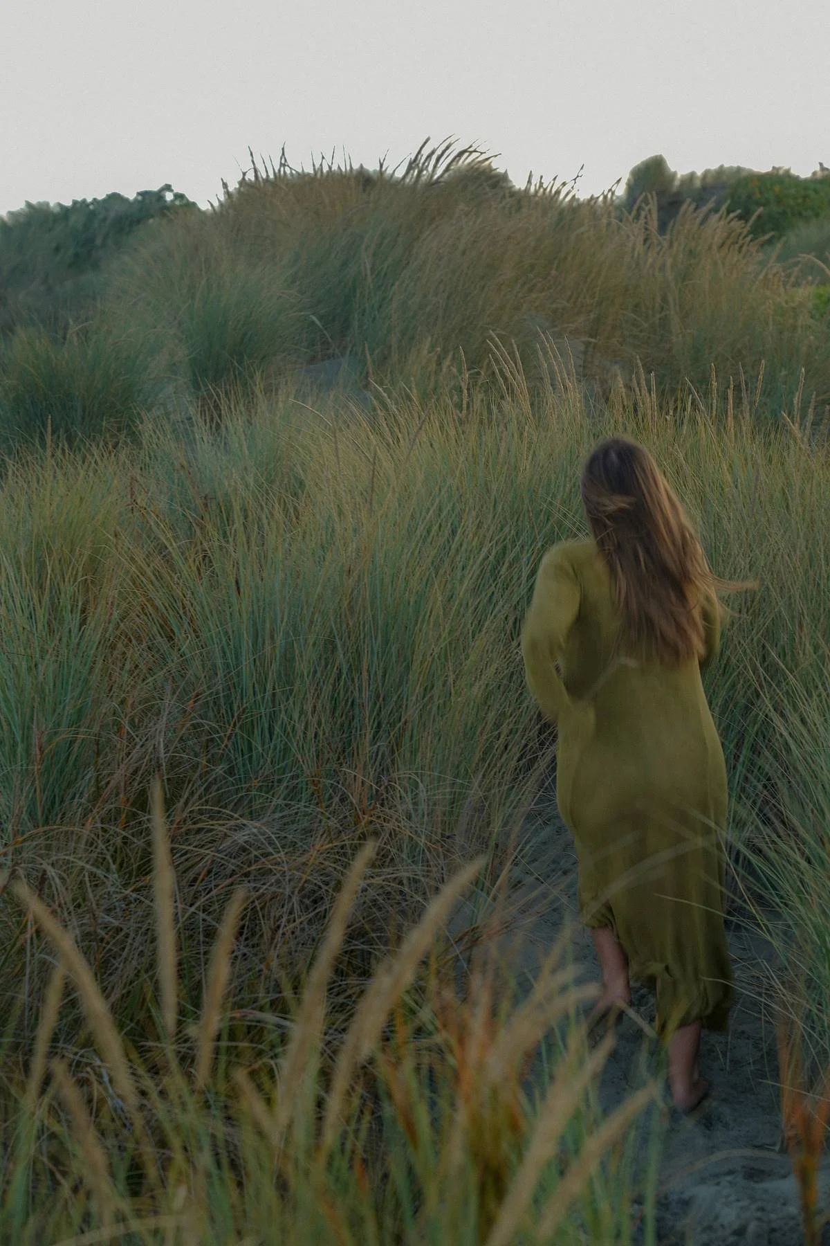 A woman with long brown hair walking through tall grass on a sandy path, wearing a yellowish-green dress, with a hilly landscape in the background under a cloudy sky.