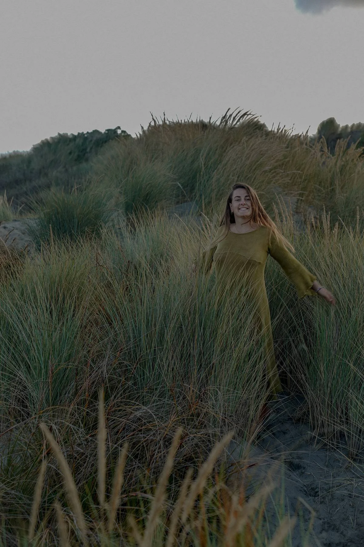 A young woman with long hair wearing a green dress smiling and walking through tall grass on a sandy terrain.