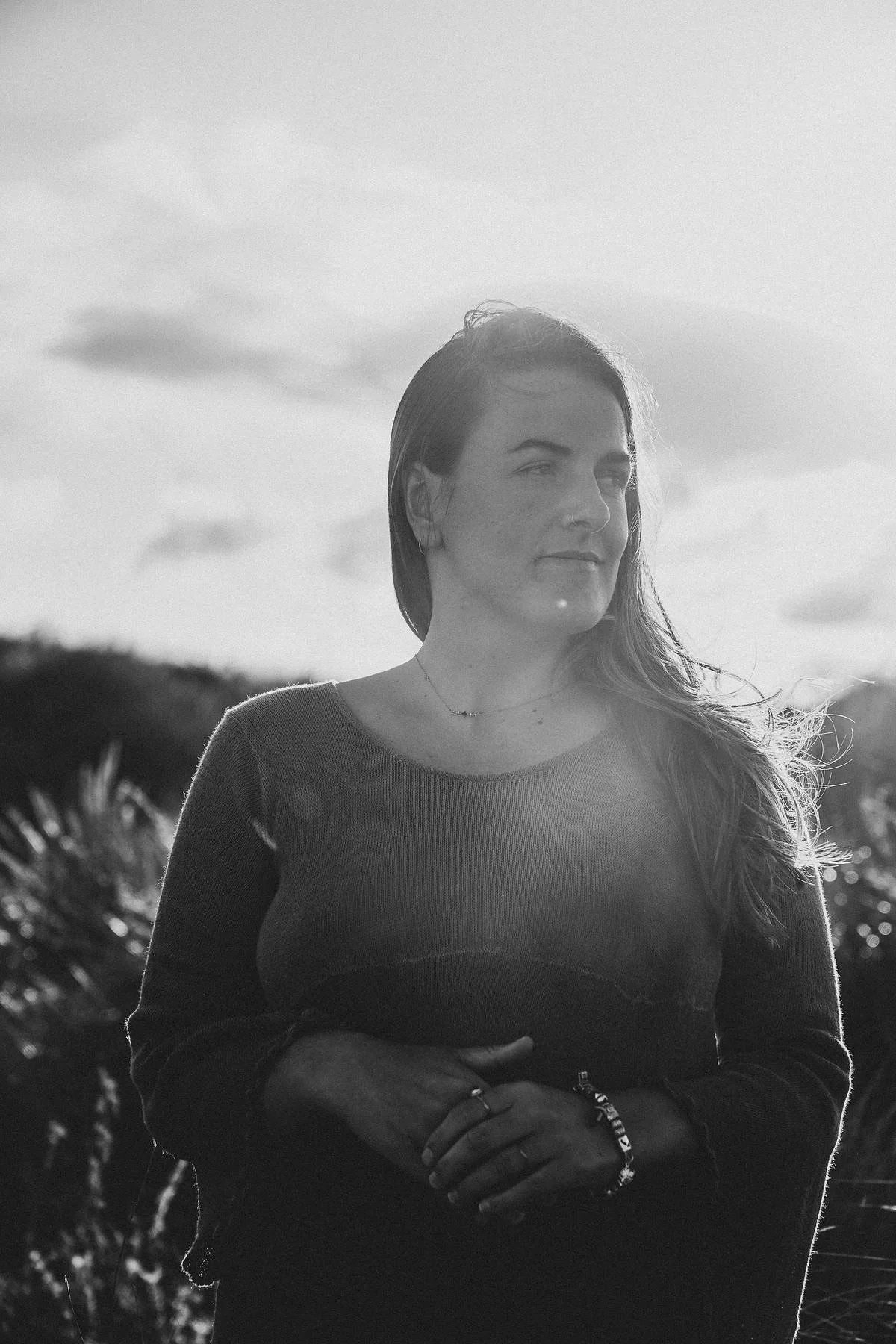 Black and white photo of a woman with long hair outdoors, with the sun behind her, trees in the background, and her hands clasped in front of her