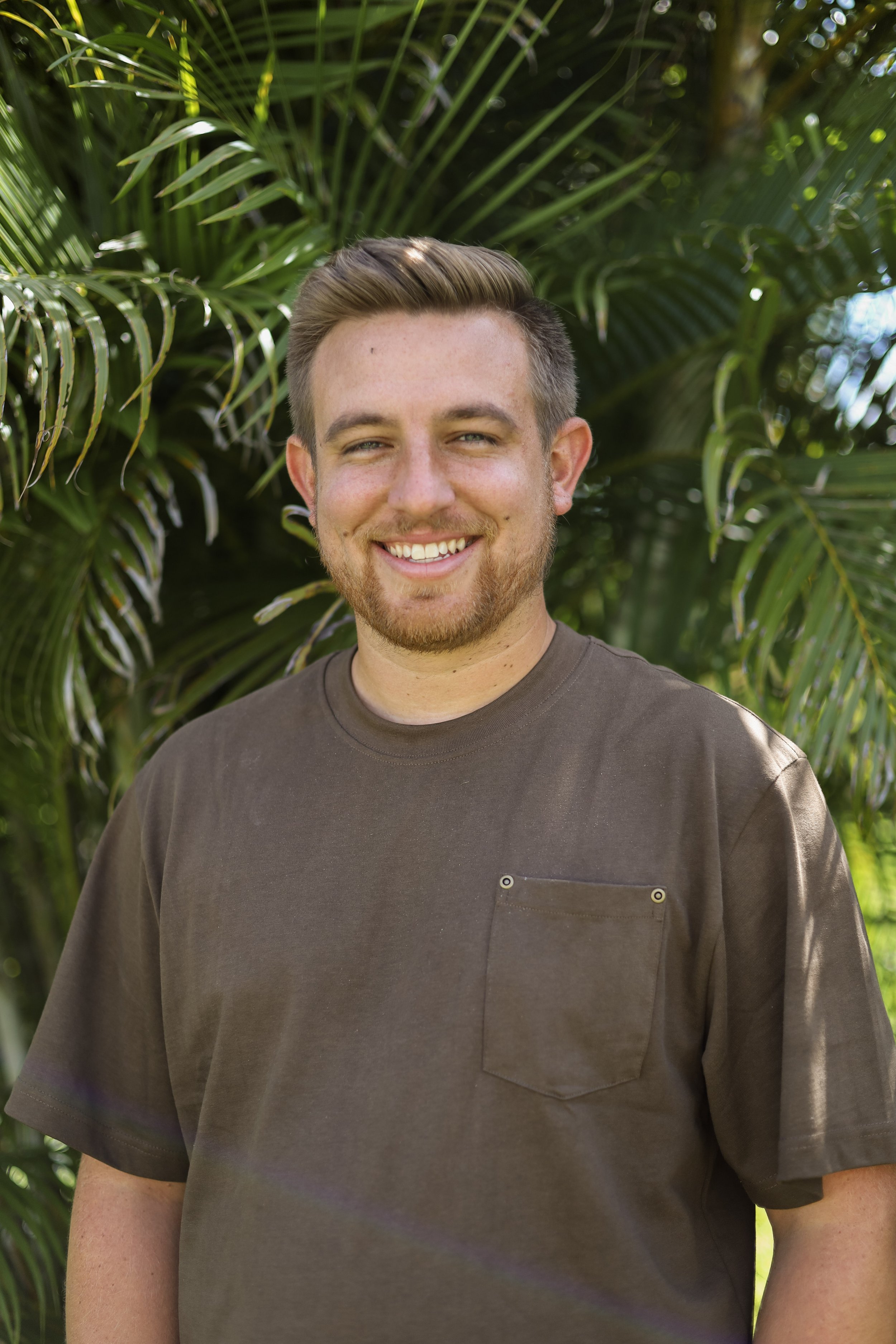 A young man with short hair, a beard, and wearing a dark brown t-shirt standing outdoors in front of lush green foliage, smiling at the camera.