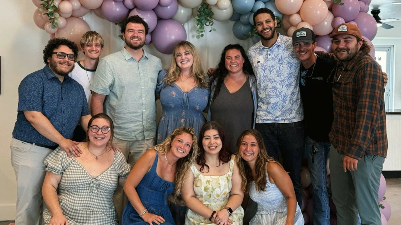 A group of fifteen people, smiling and posing together, in front of a backdrop decorated with purple, pink, and white balloons and some greenery. The group consists of men and women of various ages.