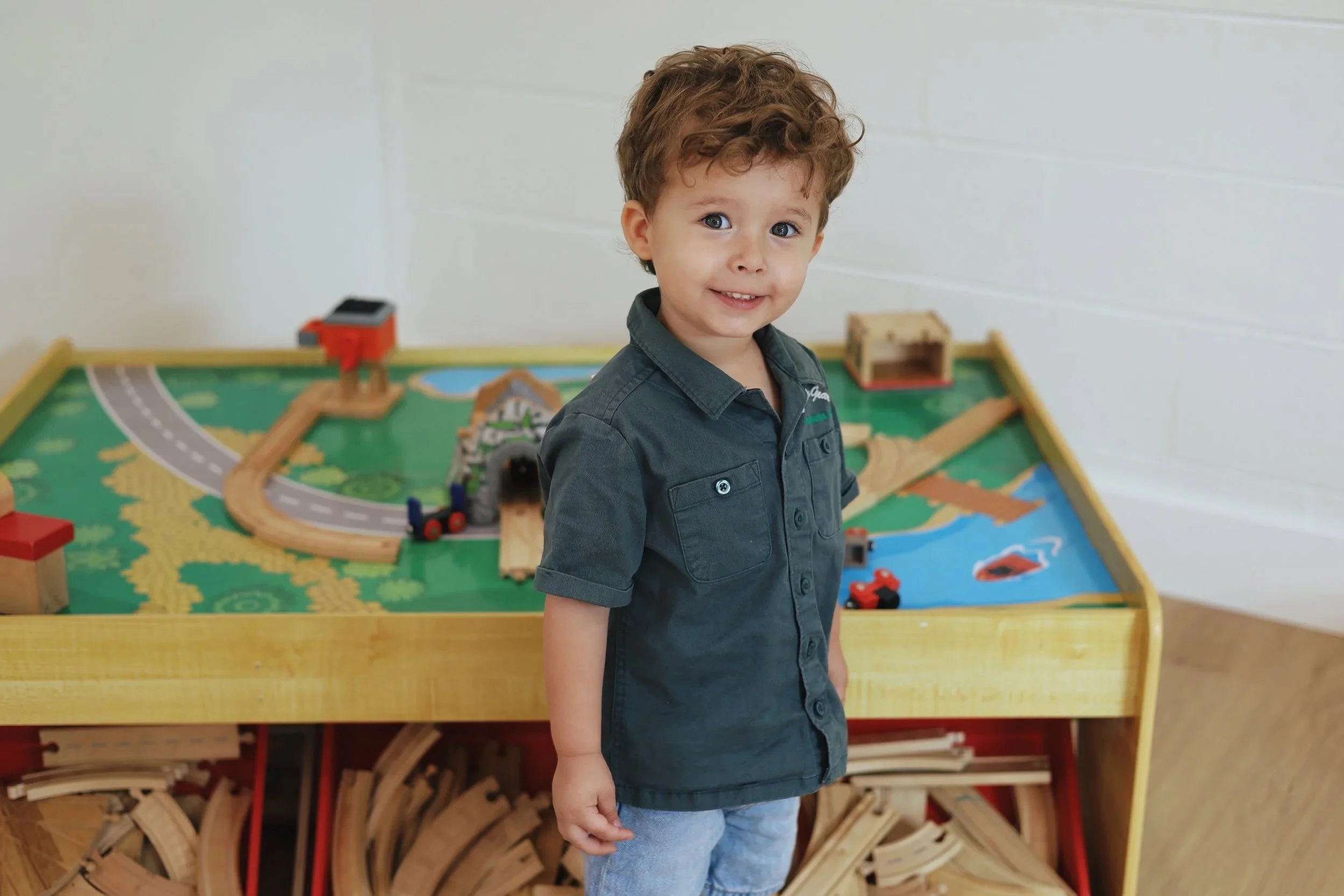 Young boy with curly hair and a blue shirt standing in front of a wooden train table with a train set. He is in the Coastline Kids preschool classroom. He loves church!