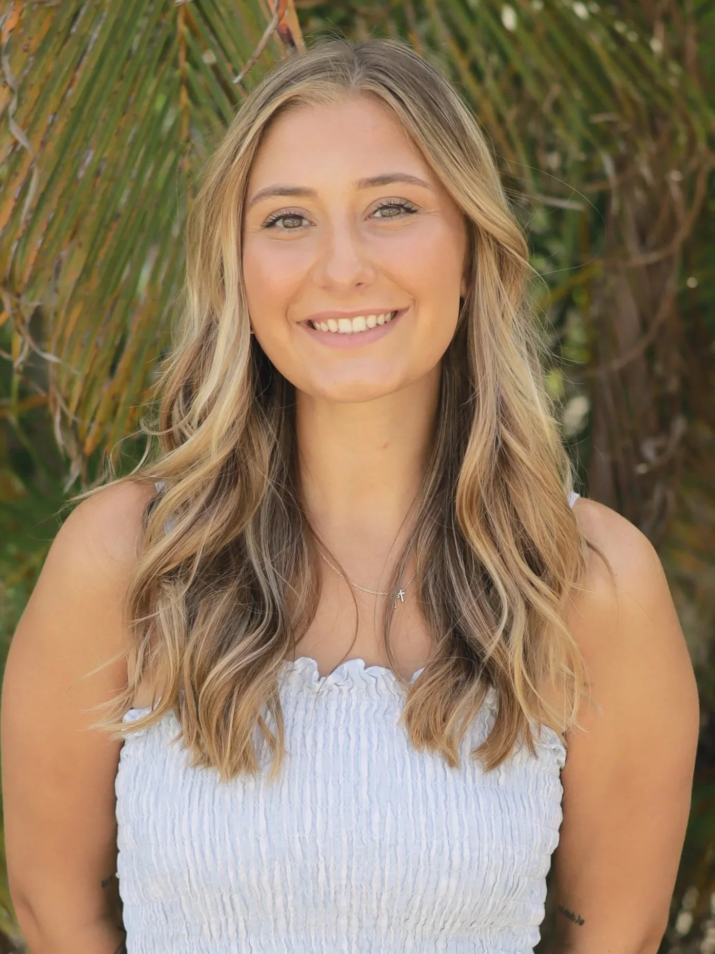 A young woman smiling outdoors in front of palm leaves, wearing a white textured sleeveless top and a delicate necklace.