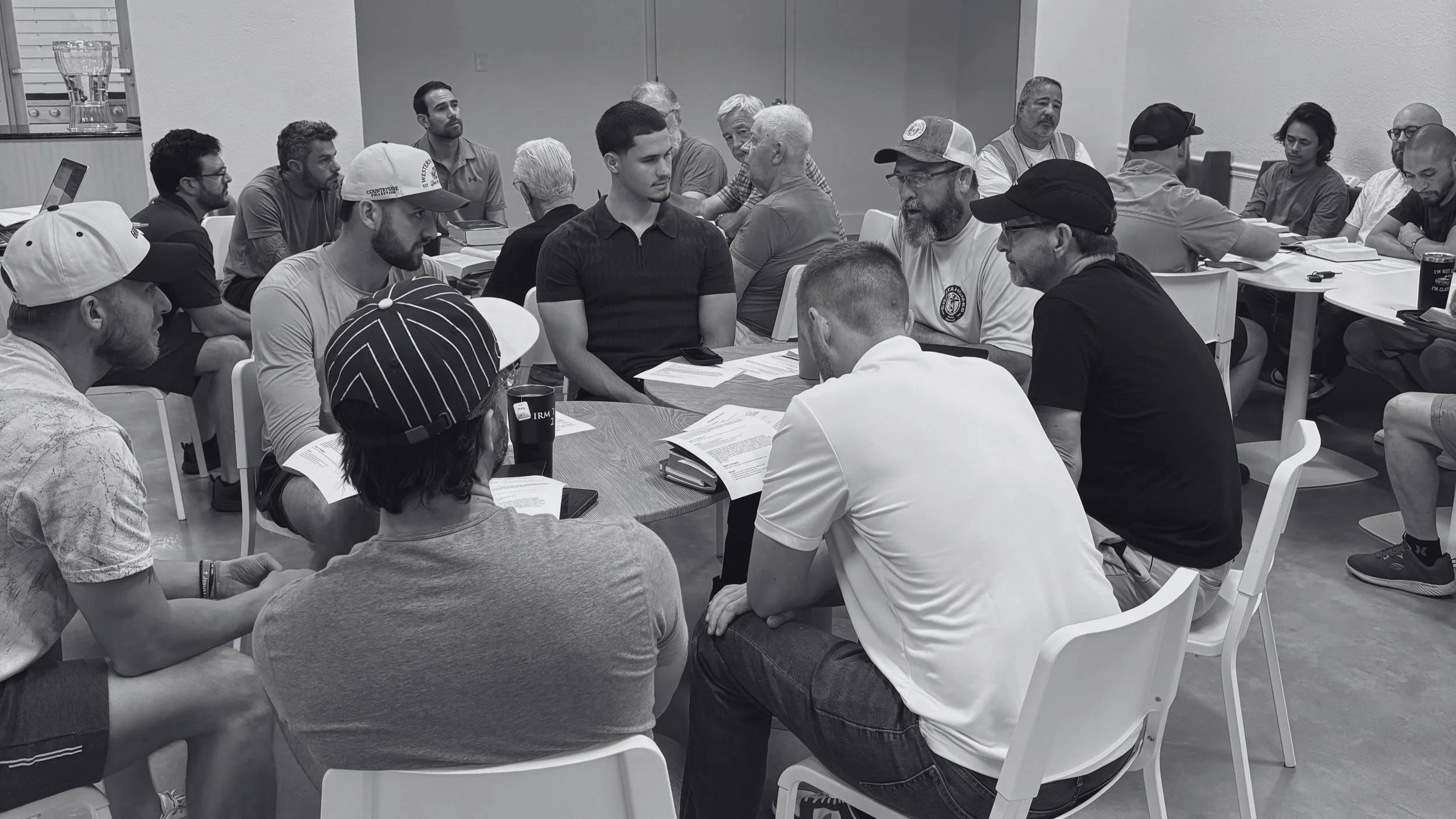 Group of people sitting around tables in a meeting room, engaged in discussion, with documents and electronic devices on the tables.