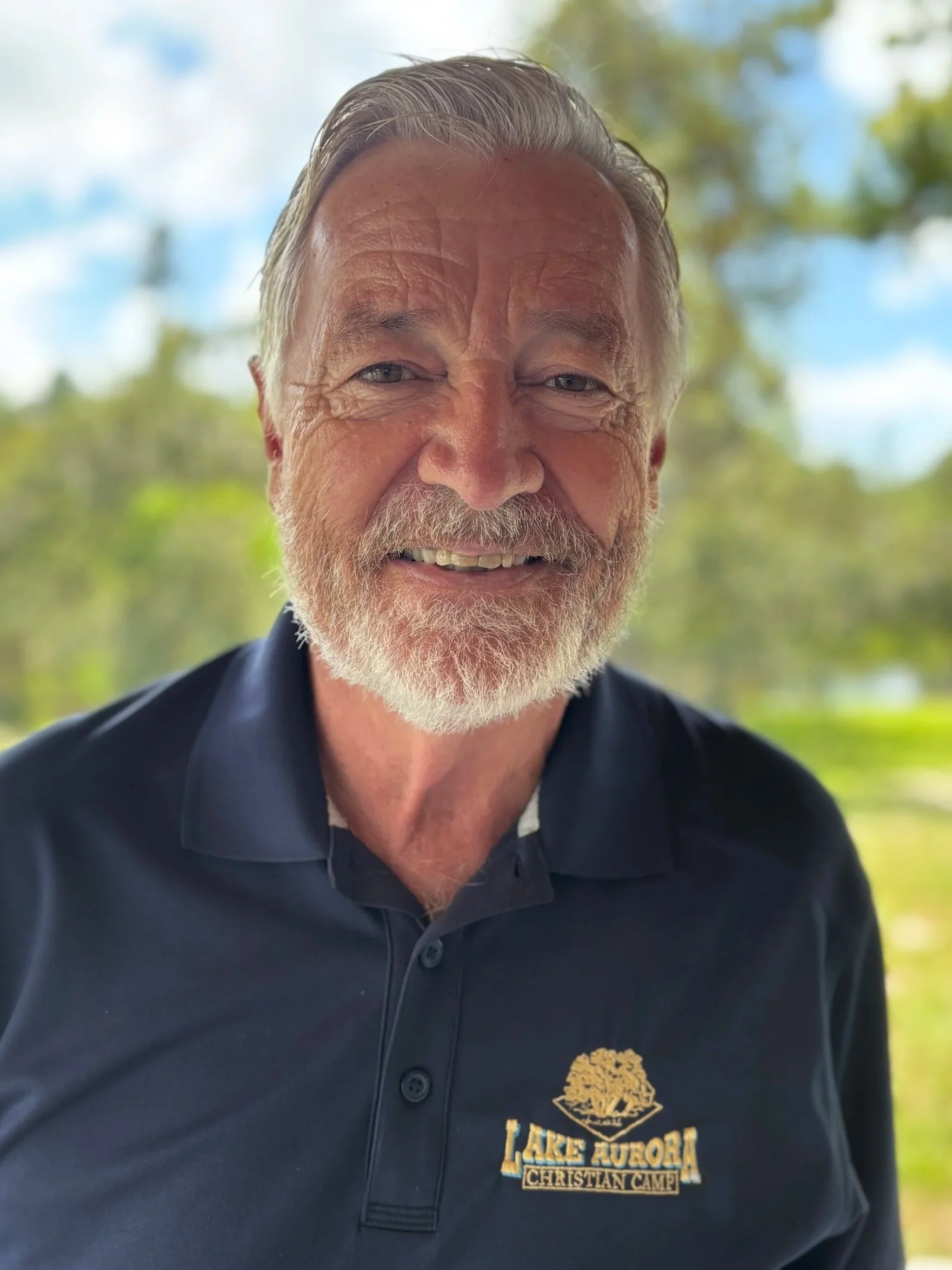 An elderly man with white hair and a beard smiling, wearing a black polo shirt with a logo that reads Lake Aurora Christian Camp, standing outdoors with a blurred green and blue background. Curt Leonard is the Care Director at Coastline in SRQ