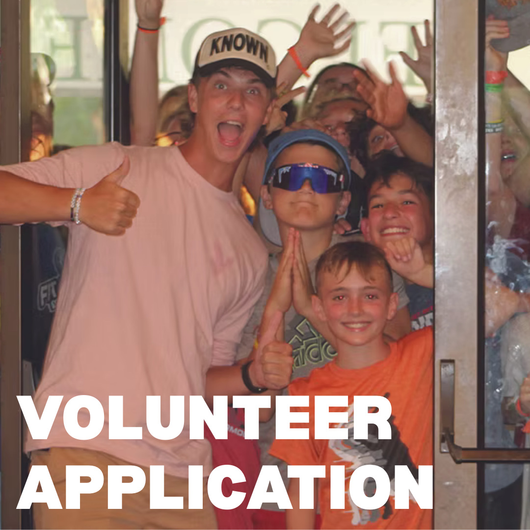 Group of excited children and teenagers excitedly pose in a doorway, some making thumbs-up signs, with a girl in a pink shirt and a boy with prayer hands, celebrating volunteering.