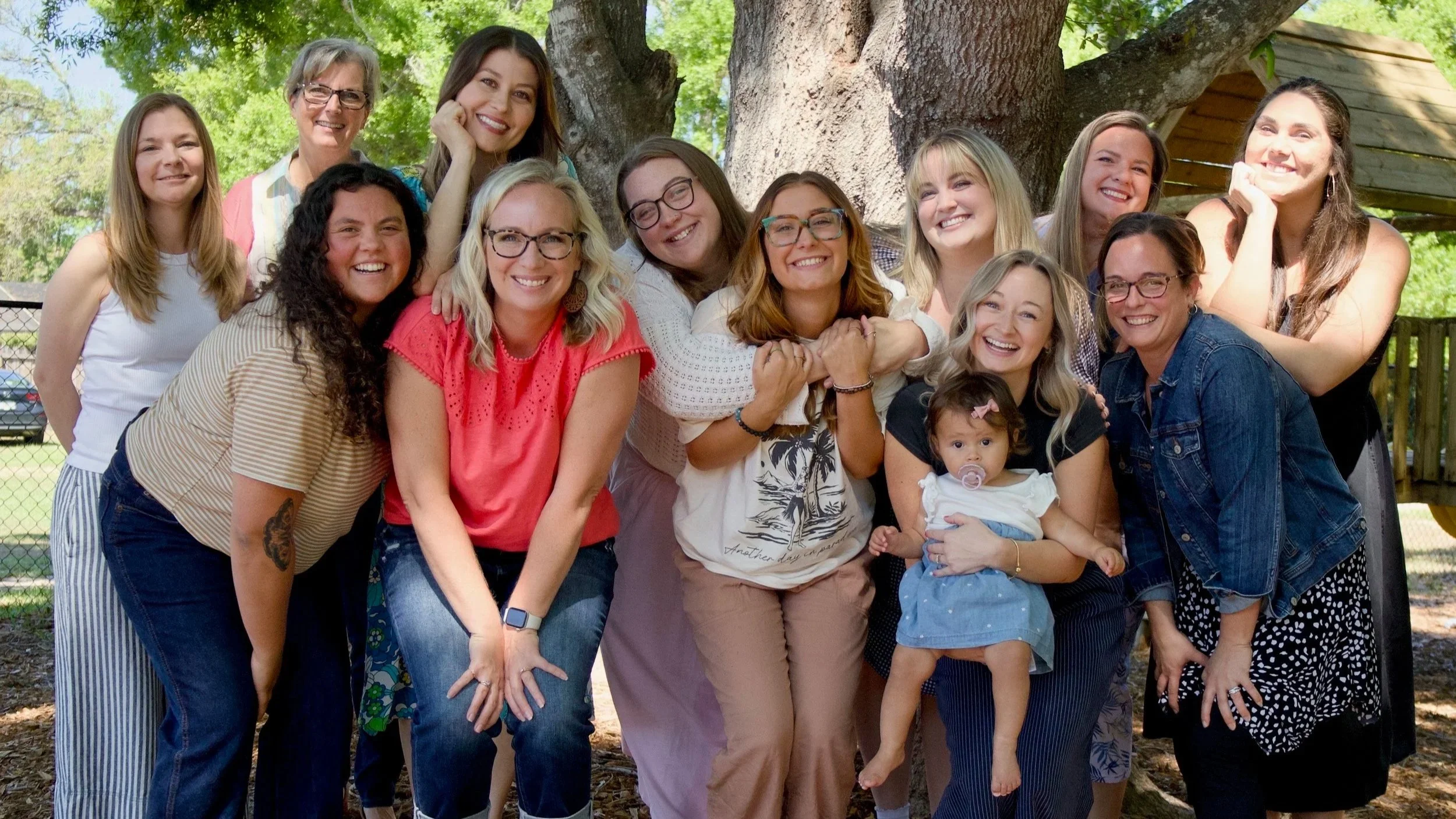 A diverse group of women and a young girl smiling outdoors with trees in the background.