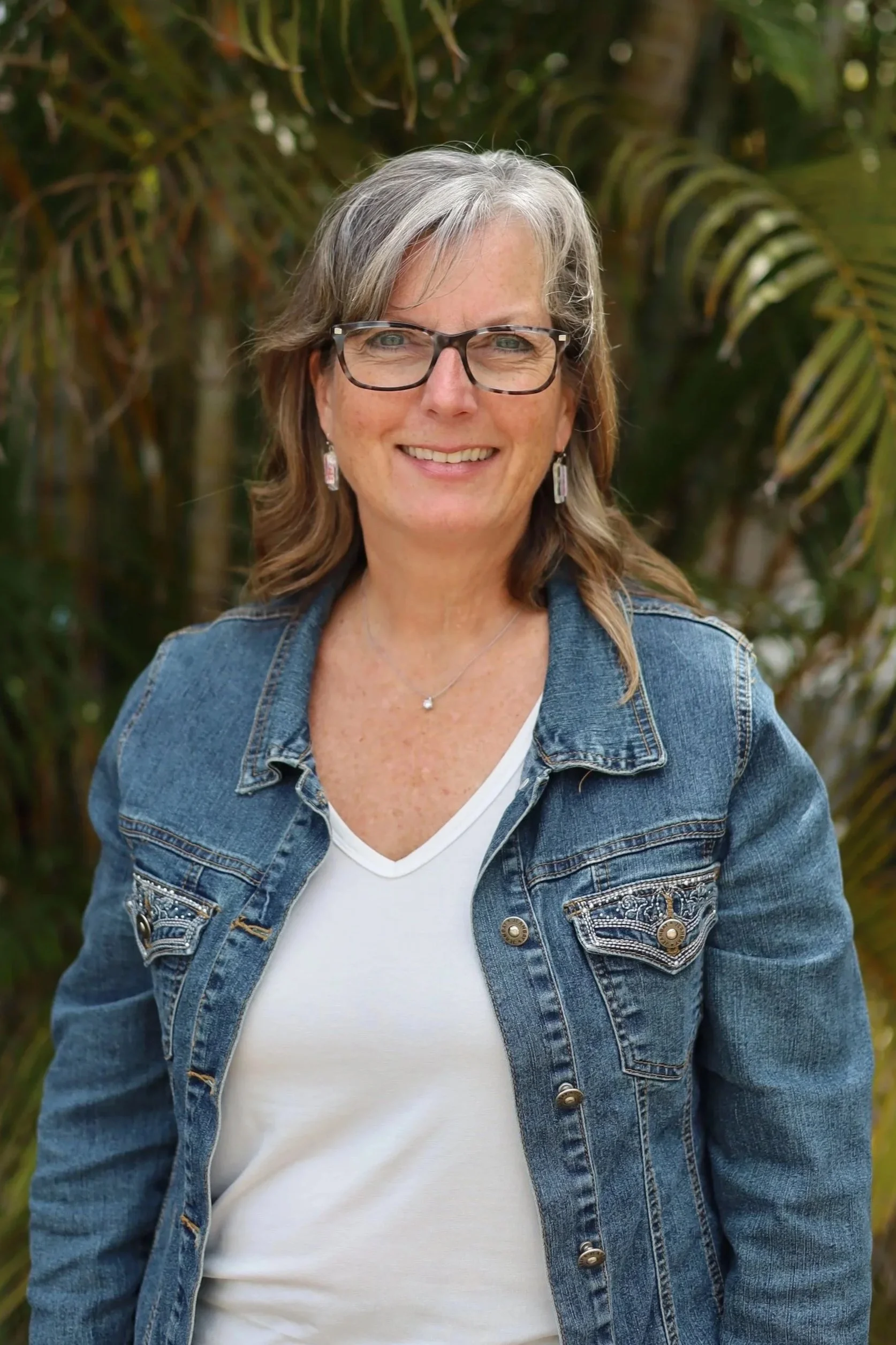 A woman with gray hair and glasses, smiling outdoors in front of greenery, wearing a denim jacket and white shirt.