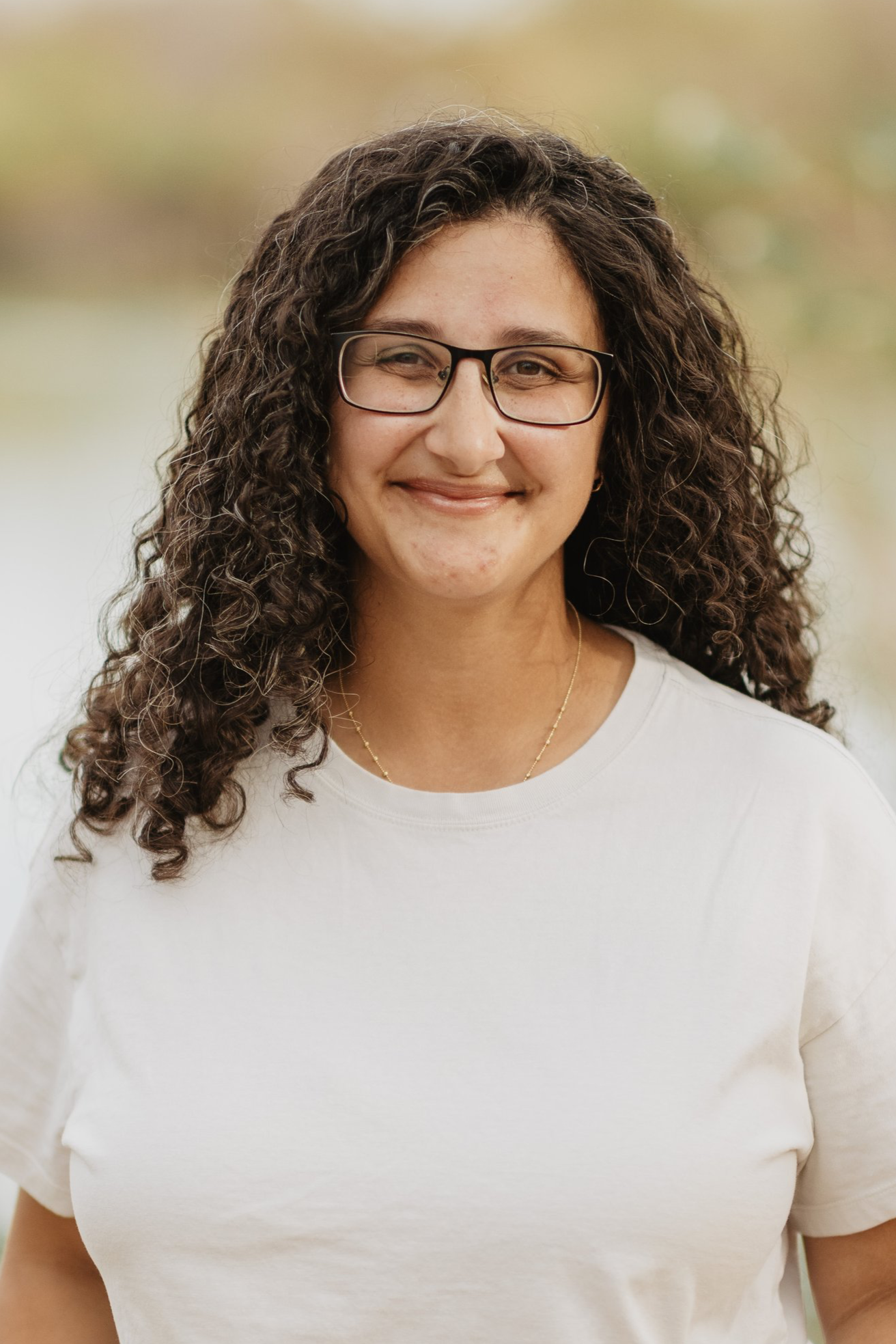 A woman with curly brown hair and glasses smiling outdoors, wearing a white t-shirt and a gold necklace. Amani is the belonging director at Coastline Christian Church in Sarasota, FL