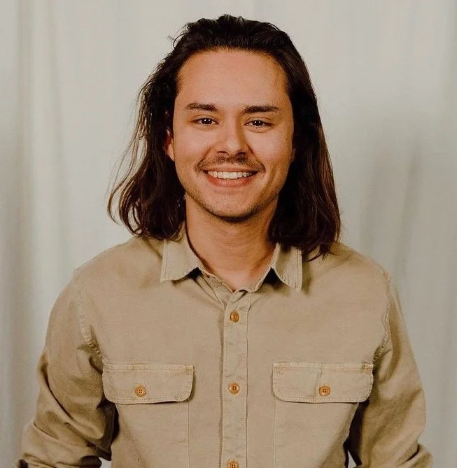 A young man with long dark hair, smiling, wearing a beige button-up shirt with pockets, standing in front of a neutral light-colored background. Brandon Simoes is the worship director at Coastline Christian Church in Sarasota