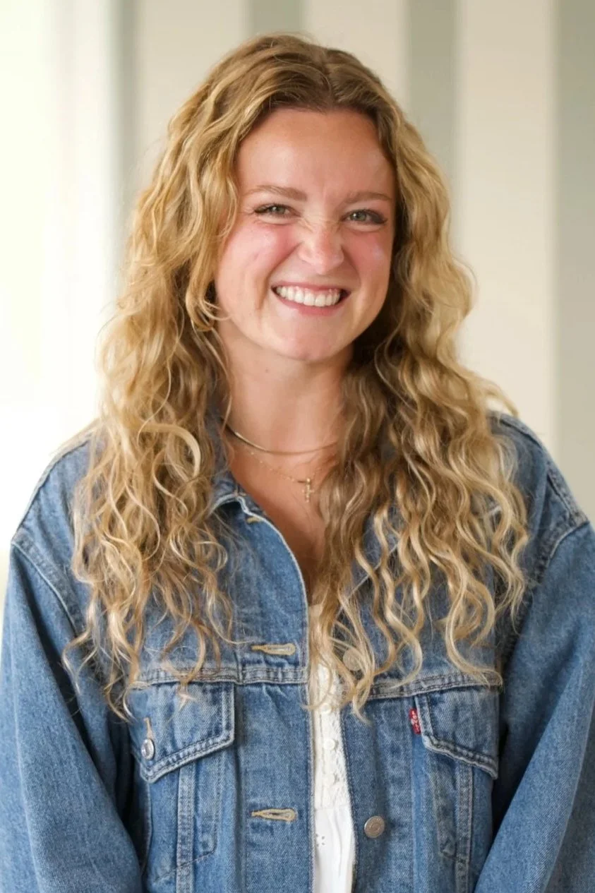 A young woman with long, curly blonde hair smiling and wearing a denim jacket over a white top, standing indoors. She is the Coastline Kids church director.