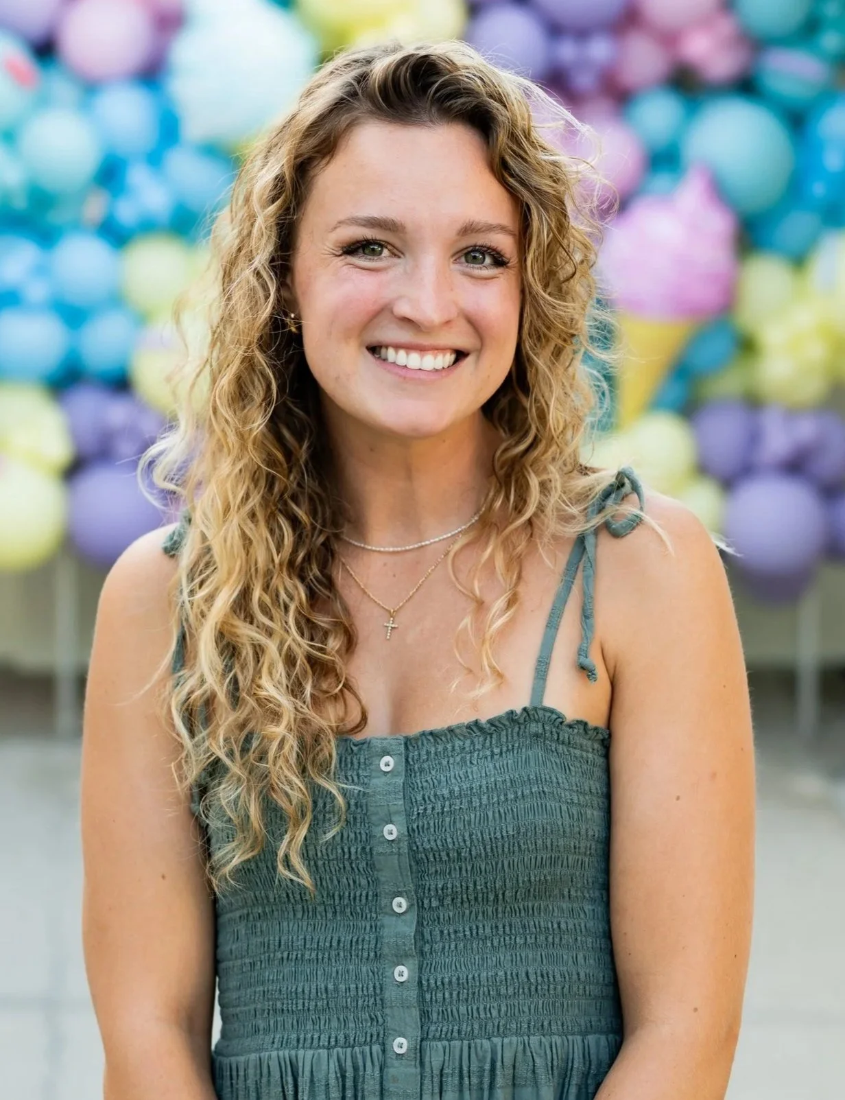 A young woman with long, curly blonde hair smiling outdoors with colorful balloons in the background.