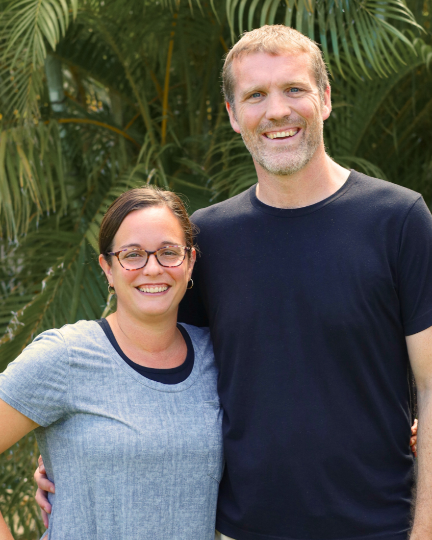A smiling woman with glasses and a man with a short beard and blonde hair standing outdoors in front of green palm trees. Michael & Kristen Elliott are the outreach directors at Coastline Christian Church in Sarasota, FL.