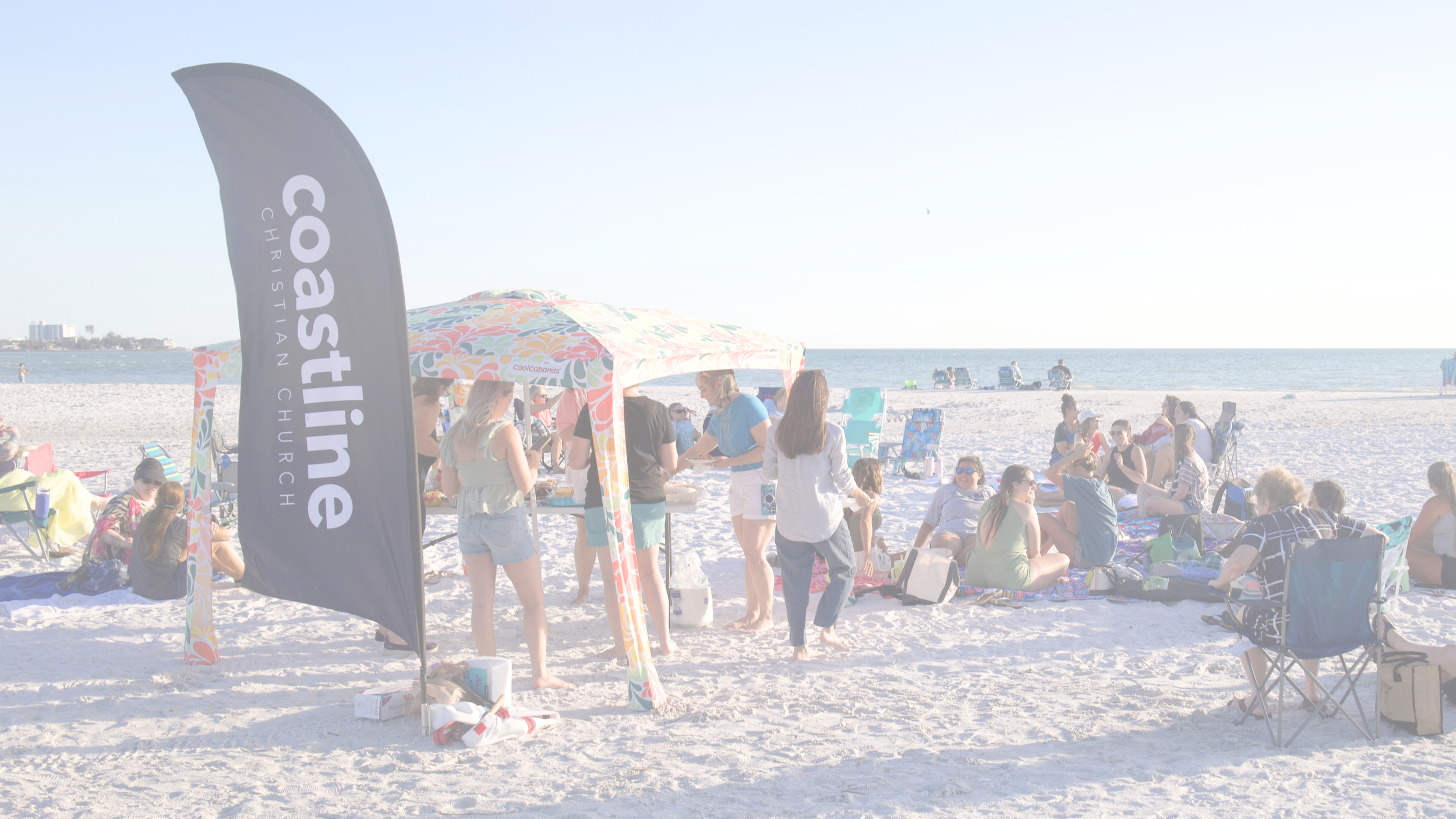 People gathered on a sandy beach, some sitting and some standing, with colorful umbrellas and a black flag with white text promoting a churro business, under a clear sky with the ocean in the background.