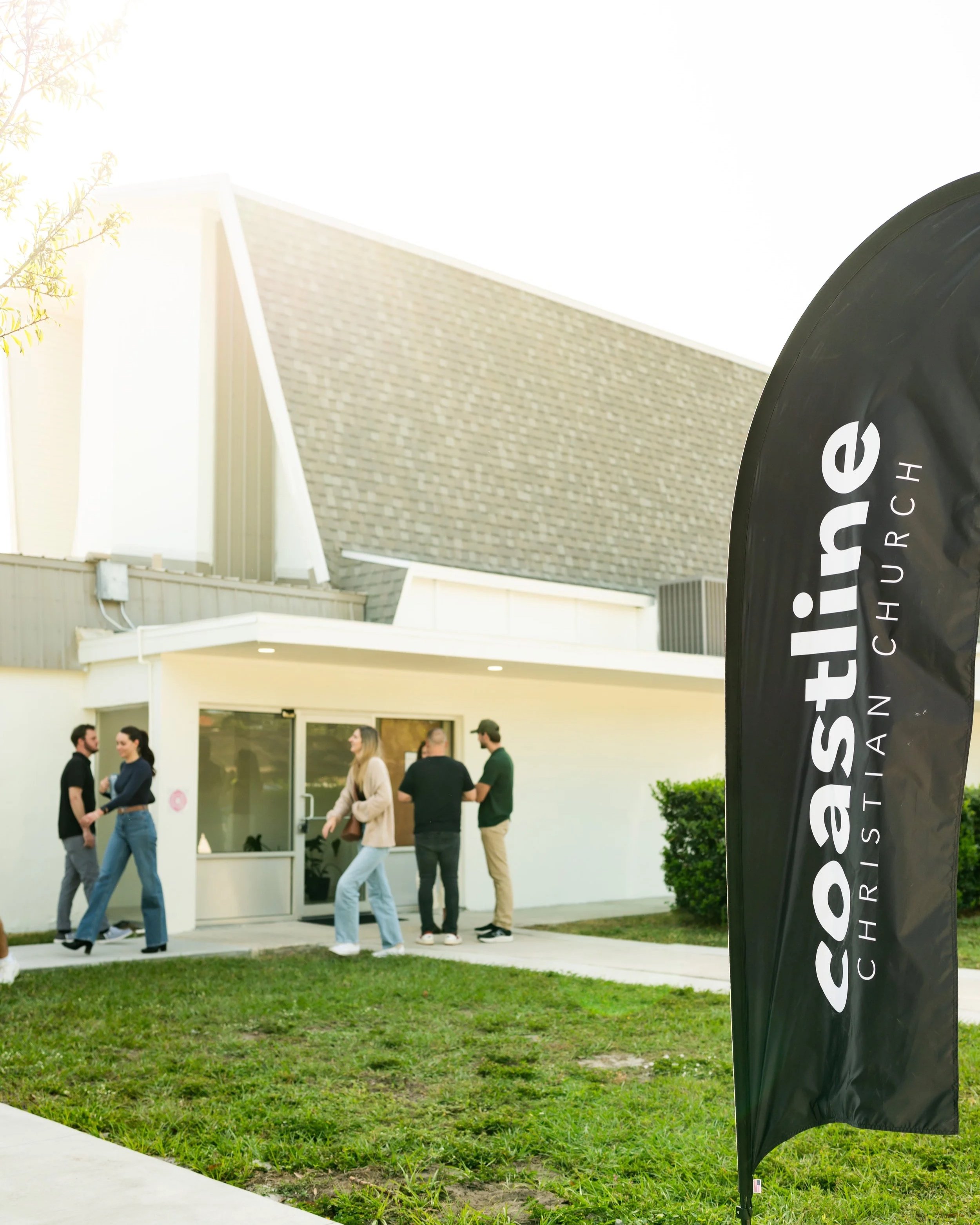 People standing and talking outside of a church in Sarasota, Florida with a black flag that says 'Coastline Christian Church' in white text.