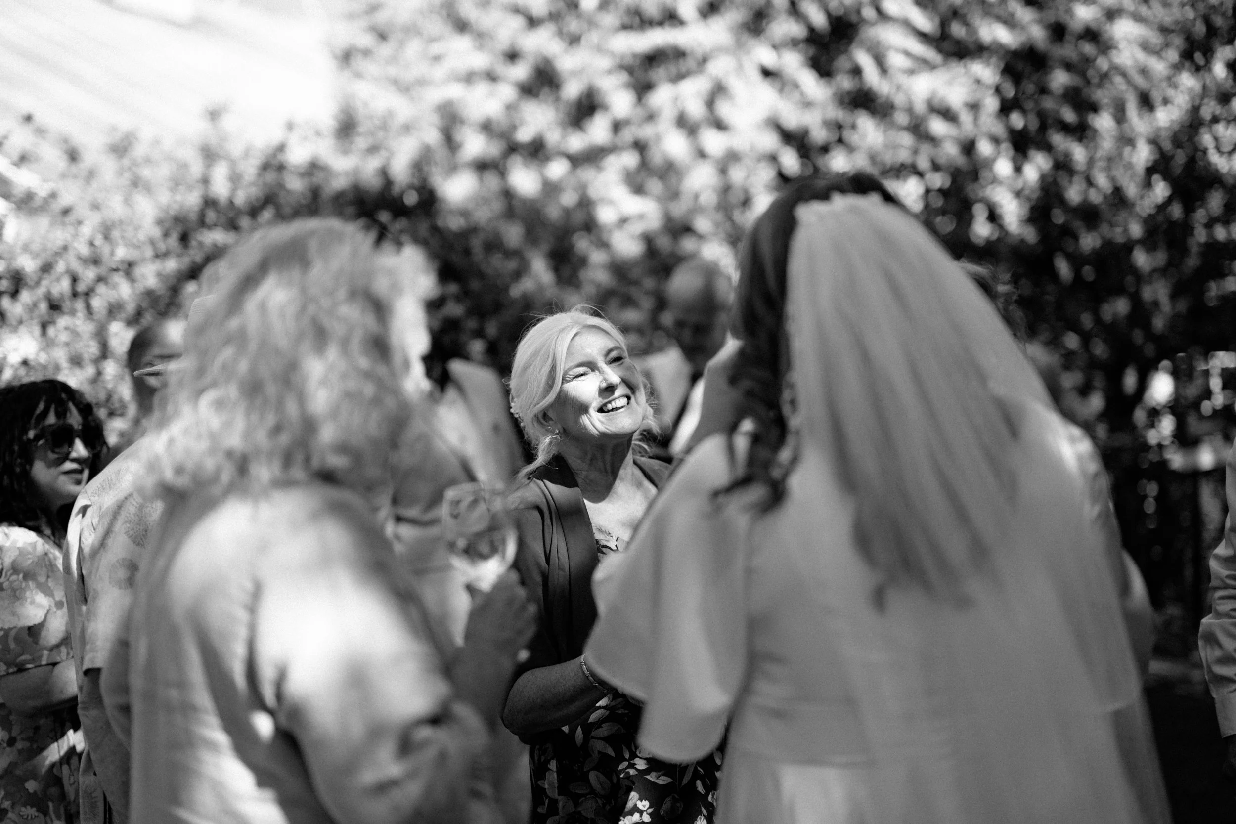 Kaiapoi Wedding, Canterbury, New Zealand. A smiling woman talking to bride in a wedding dress, surrounded by other guests, outdoors with trees in the background.