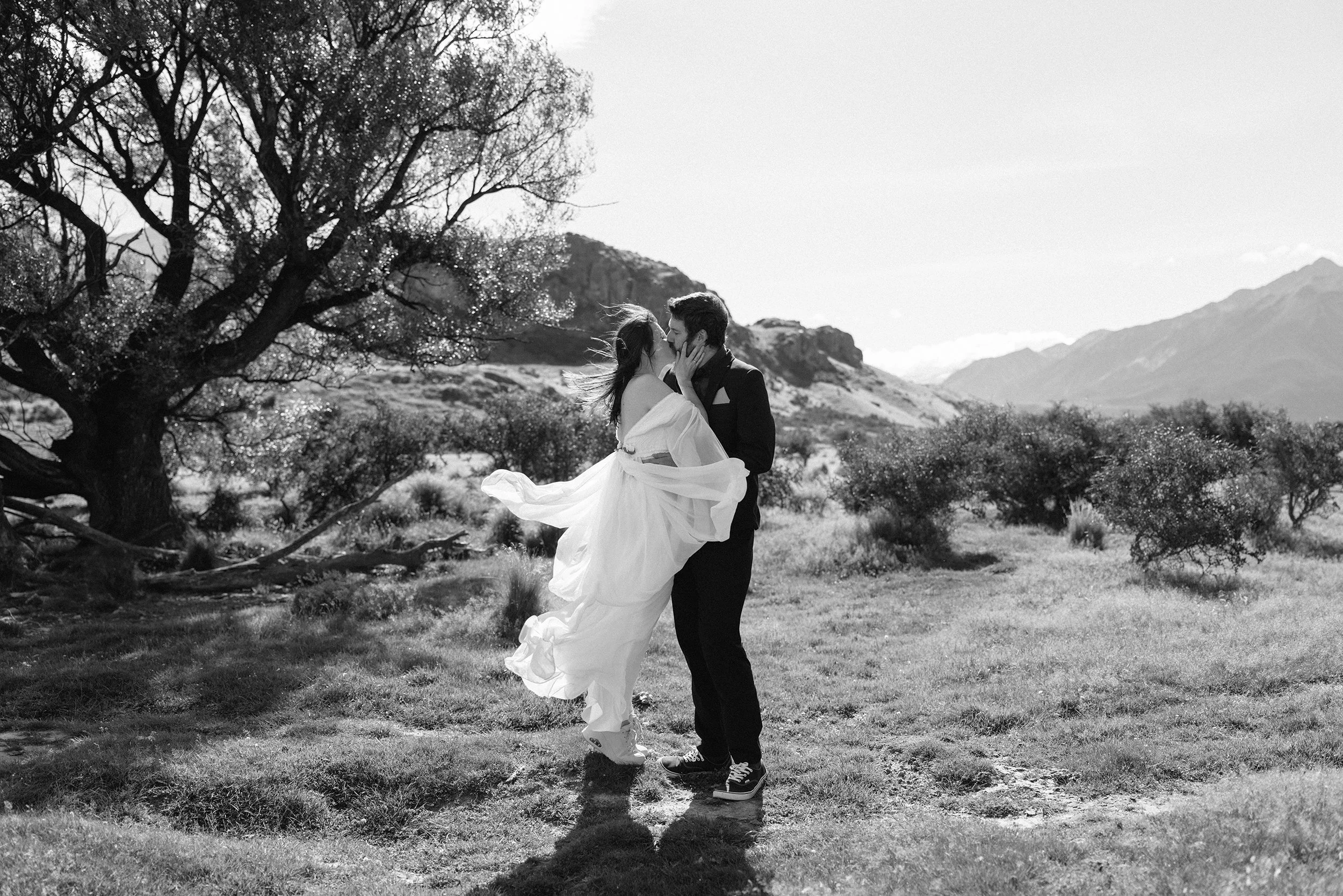 Film + Digital Wedding Photographer Christchurch - Mount Sunday Elopement, Canterbury, New Zealand. Black and white photo of bride and groom kissing with trees and mountains in the background. Bride wears a flowy white wedding dress.