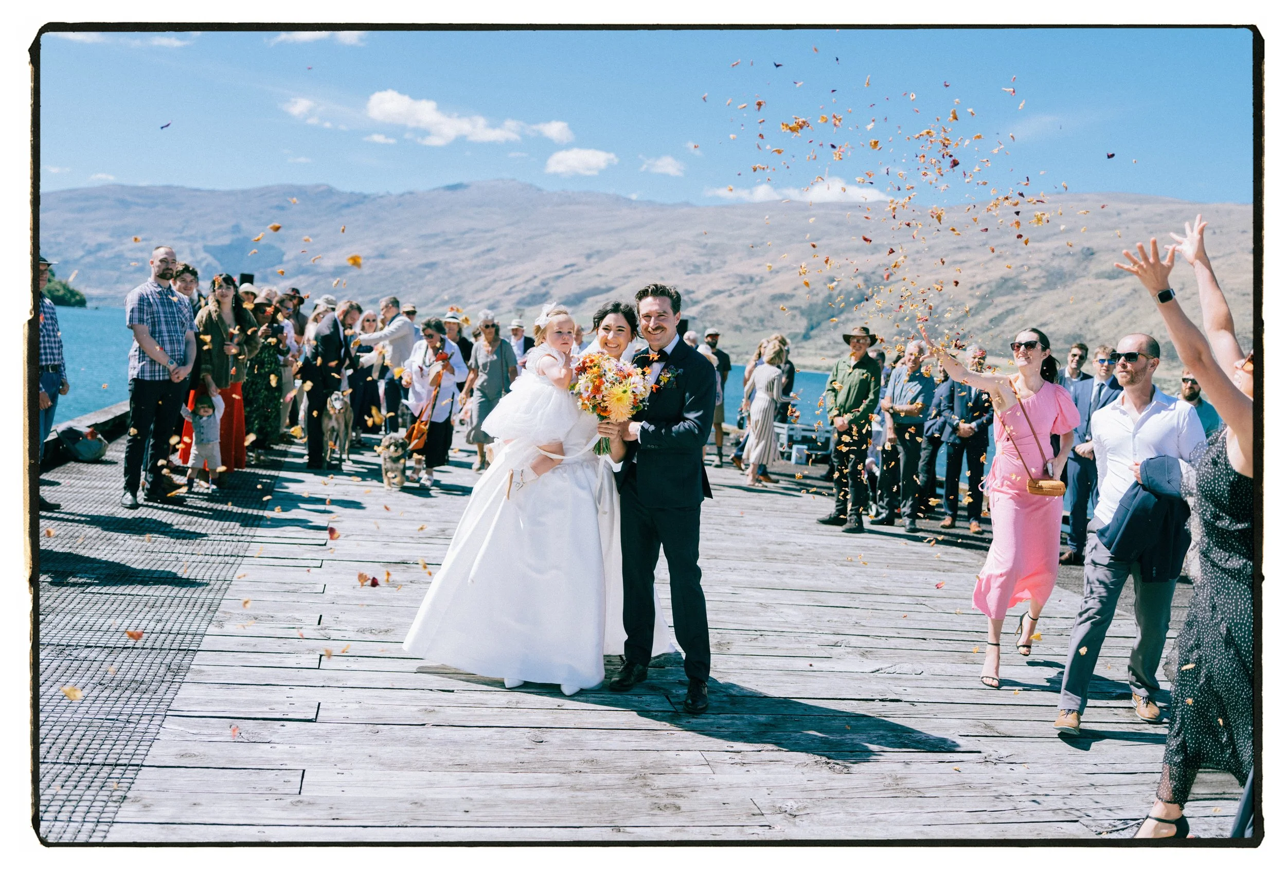 Film + Digital Wedding Photographer Christchurch - Kingston Wedding, Otago, New Zealand. Bride and groom smiling and holding each other at wedding ceremony on Wakatipu lake, with guests throwing flower petals and mountains in the background.