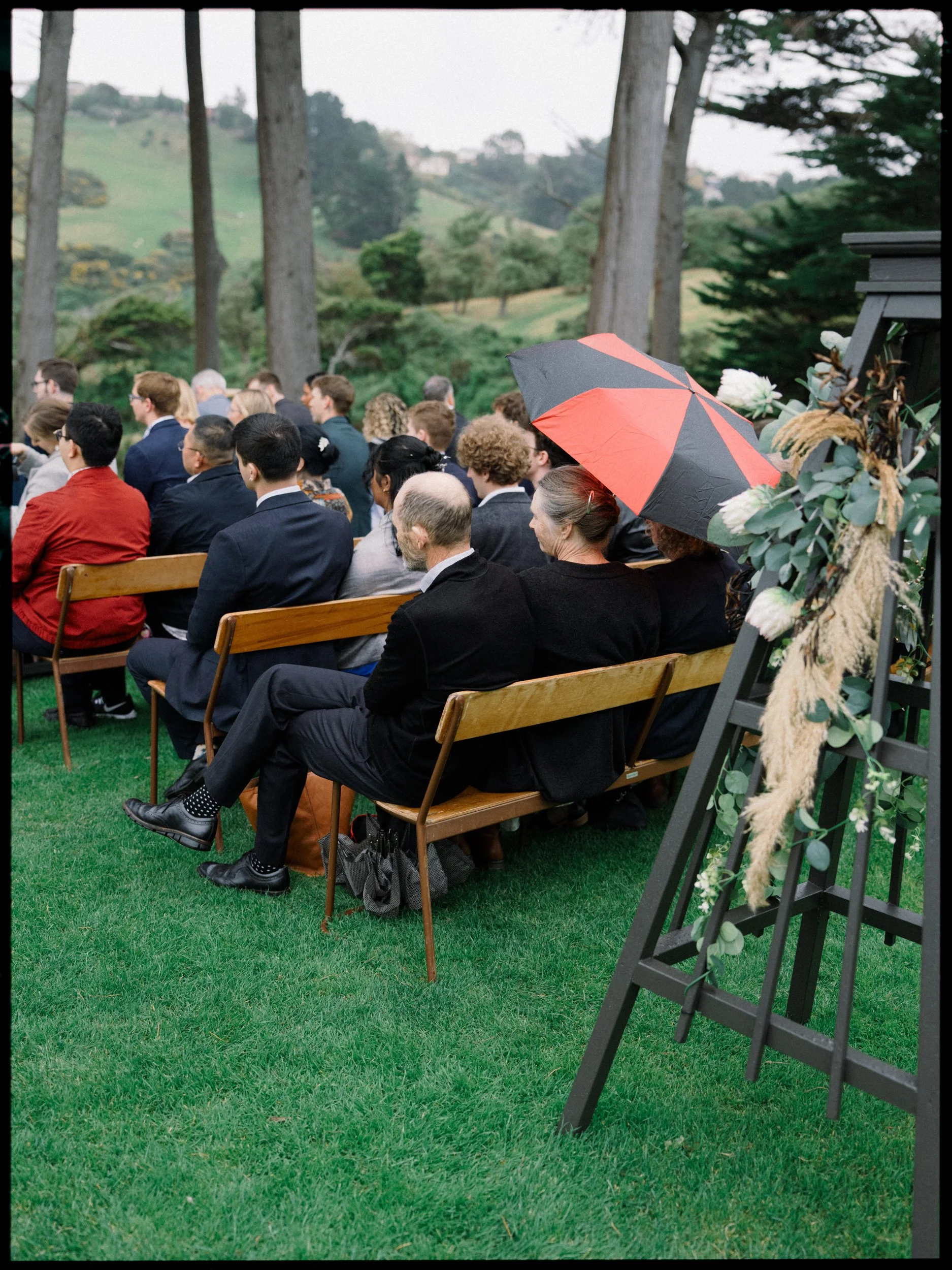 Film + Digital Wedding Photographer Dunedin - Lochend Woolshed, Dunedin, Otago, New Zealand. Guests seated outdoors during rainy wedding ceremony with woman holding a red and black umbrella surrounded by tall trees and a hilly landscape.