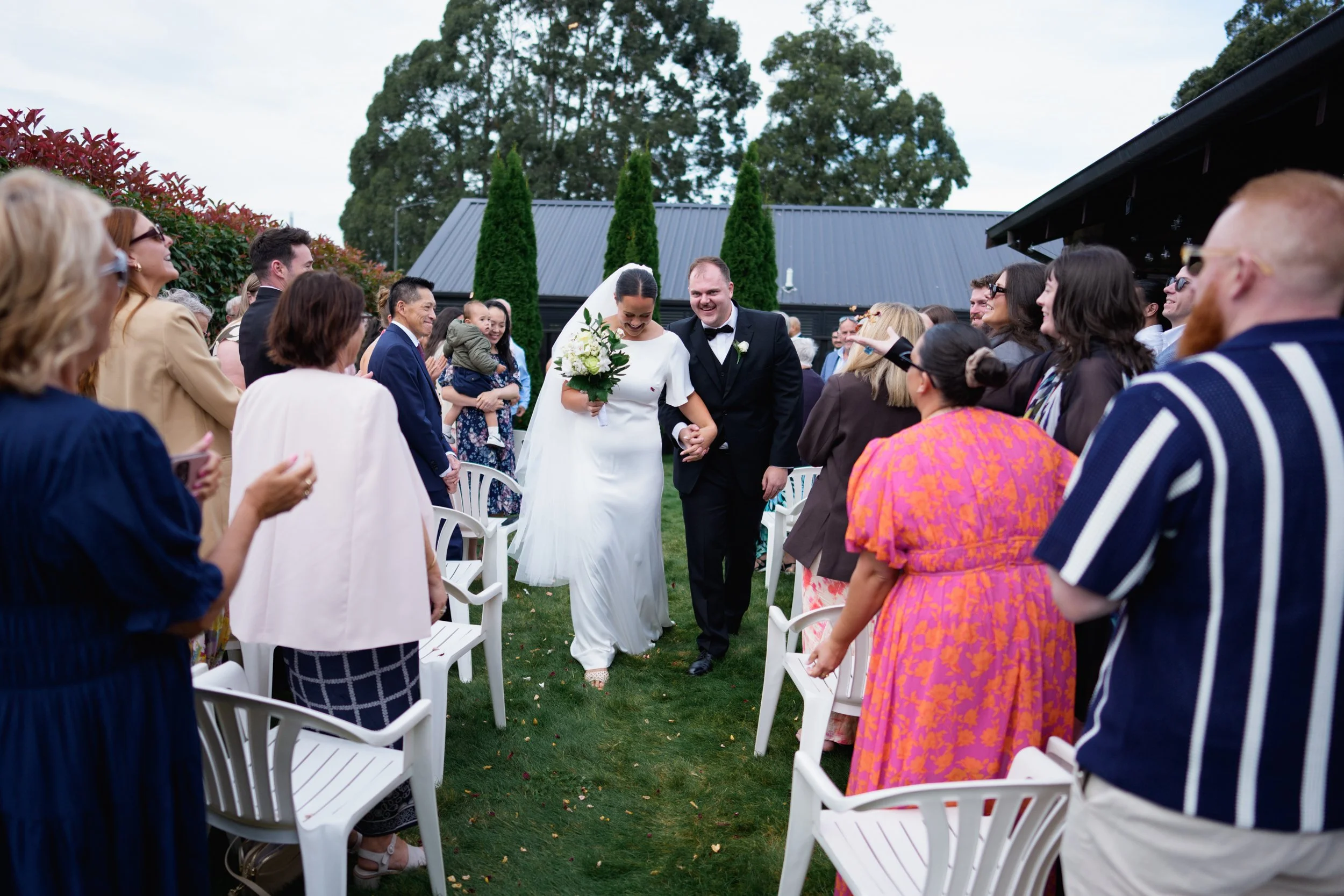 Film + Digital Wedding Photographer Christchurch - Backyard Wedding, Christchurch, Canterbury, New Zealand. Bride and groom walking down the aisle hand in hand at backyard wedding ceremony with guests celebrating.