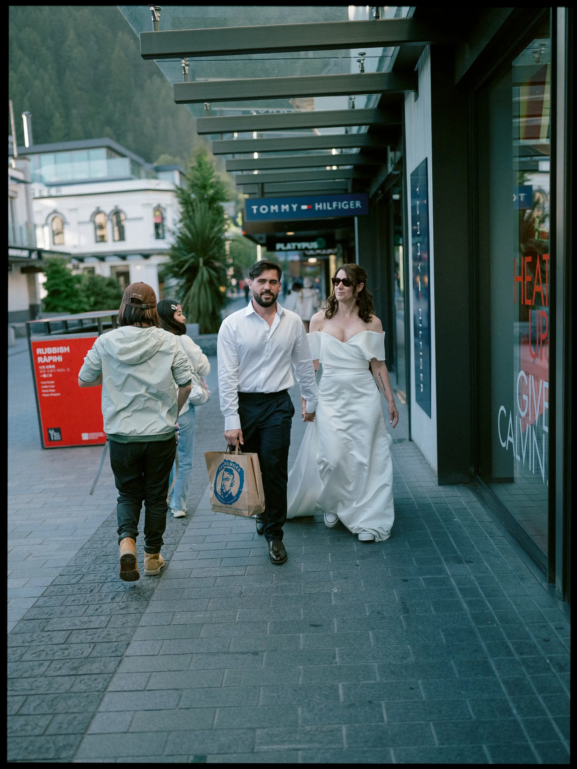 Queenstown Elopement, Otago, New Zealand. Bride in wedding dress and sunglasses and groom holding hands and walking down the street in Queenstown, carrying a Fergburger bag, with other pedestrians around and storefronts in the background.