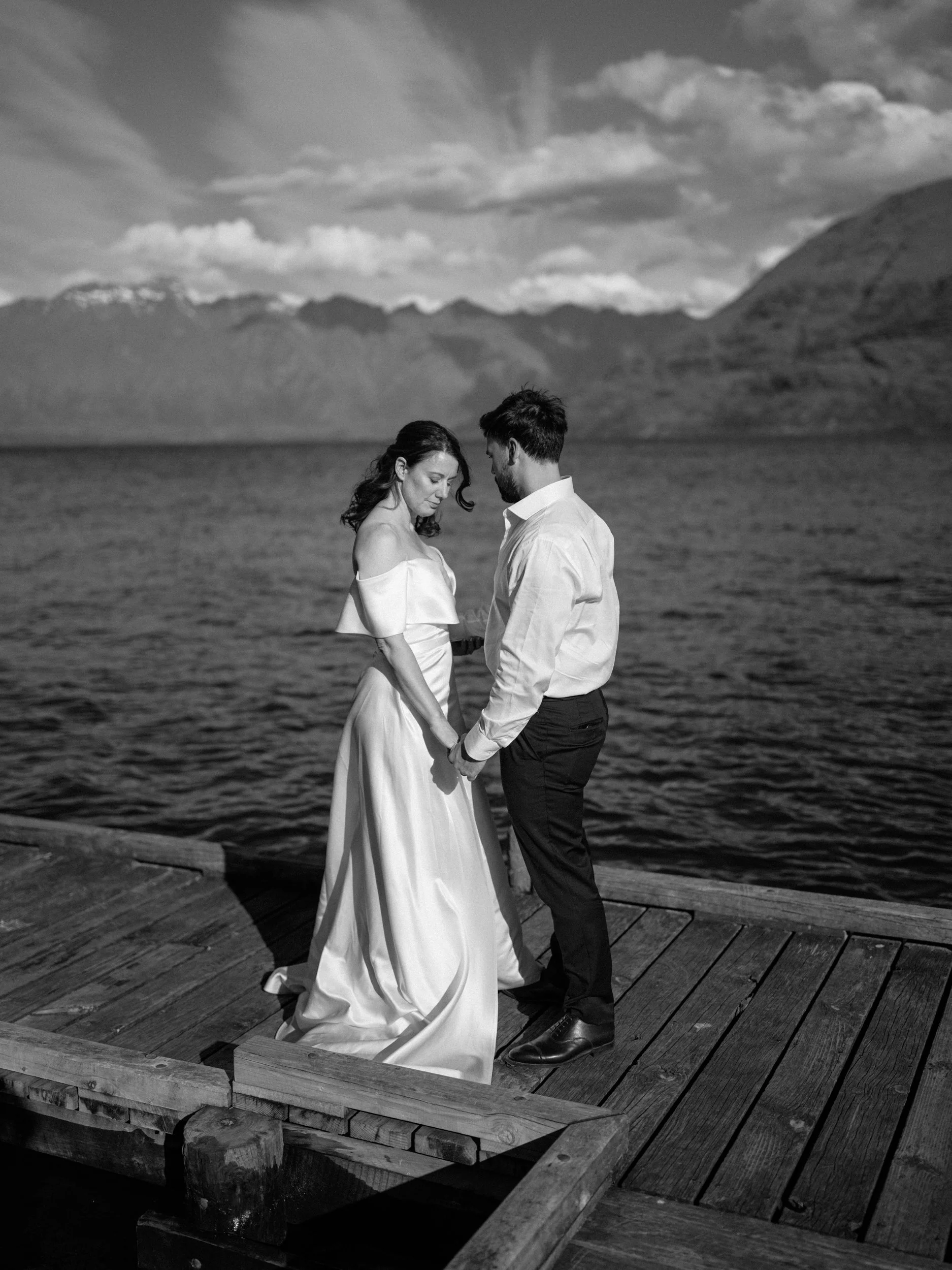 Queenstown Elopement, Otago, New Zealand. A black and white photo of bride and groom holding hands on a wooden dock by lake Wakatipu, with mountains and a cloudy sky in the background.