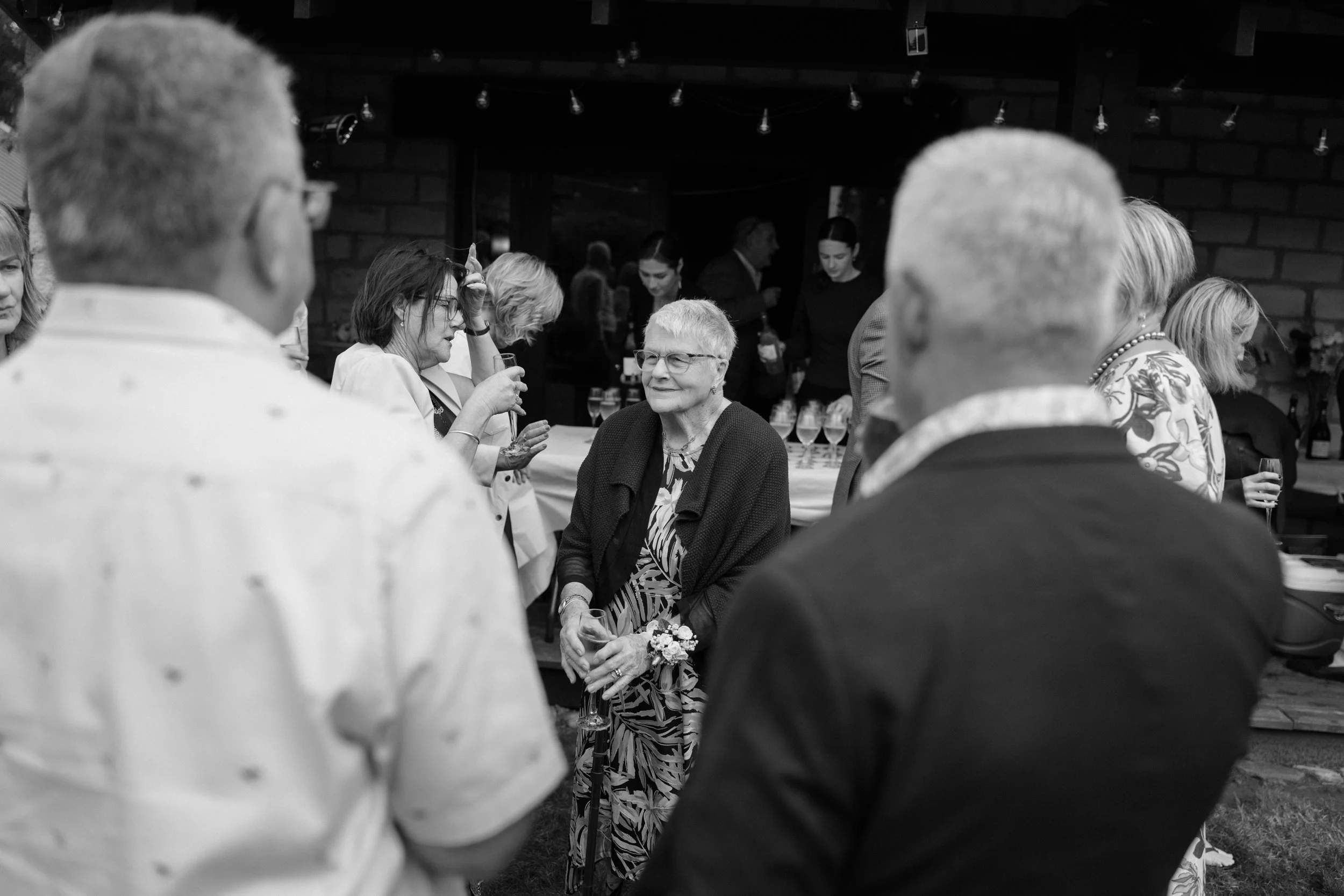 Film + Digital Wedding Photographer Christchurch - Backyard Wedding, Christchurch, Canterbury, New Zealand. Black-and-white photo of guests at backyard wedding reception, with an elderly woman in the middle.