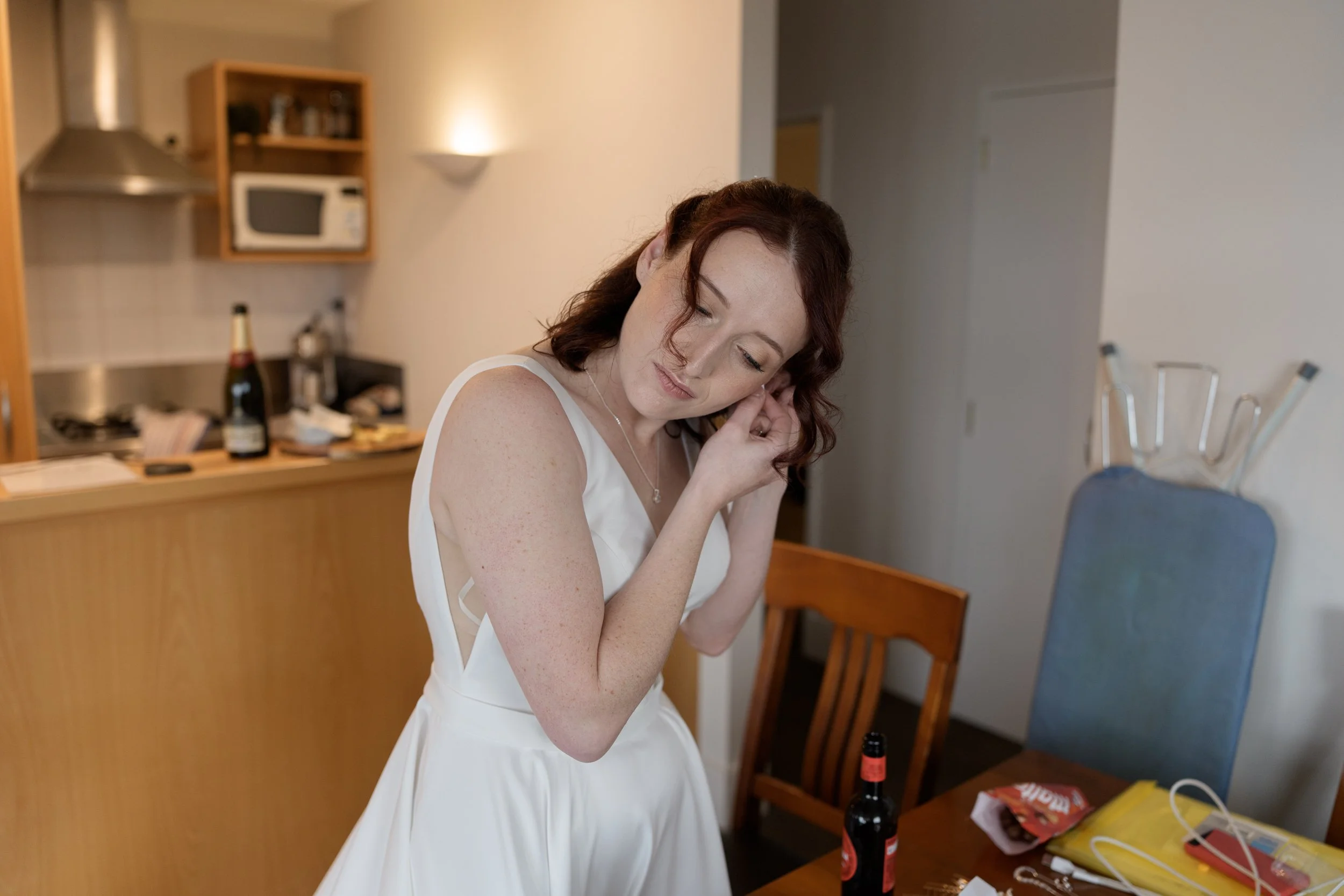 Film + Digital Wedding Photographer Queenstown - Kāmana Lakehouse, Queenstown, Otago, New Zealand. Bride dressed in a white wedding gown, adjusting her earring in a room with a kitchen in the background.