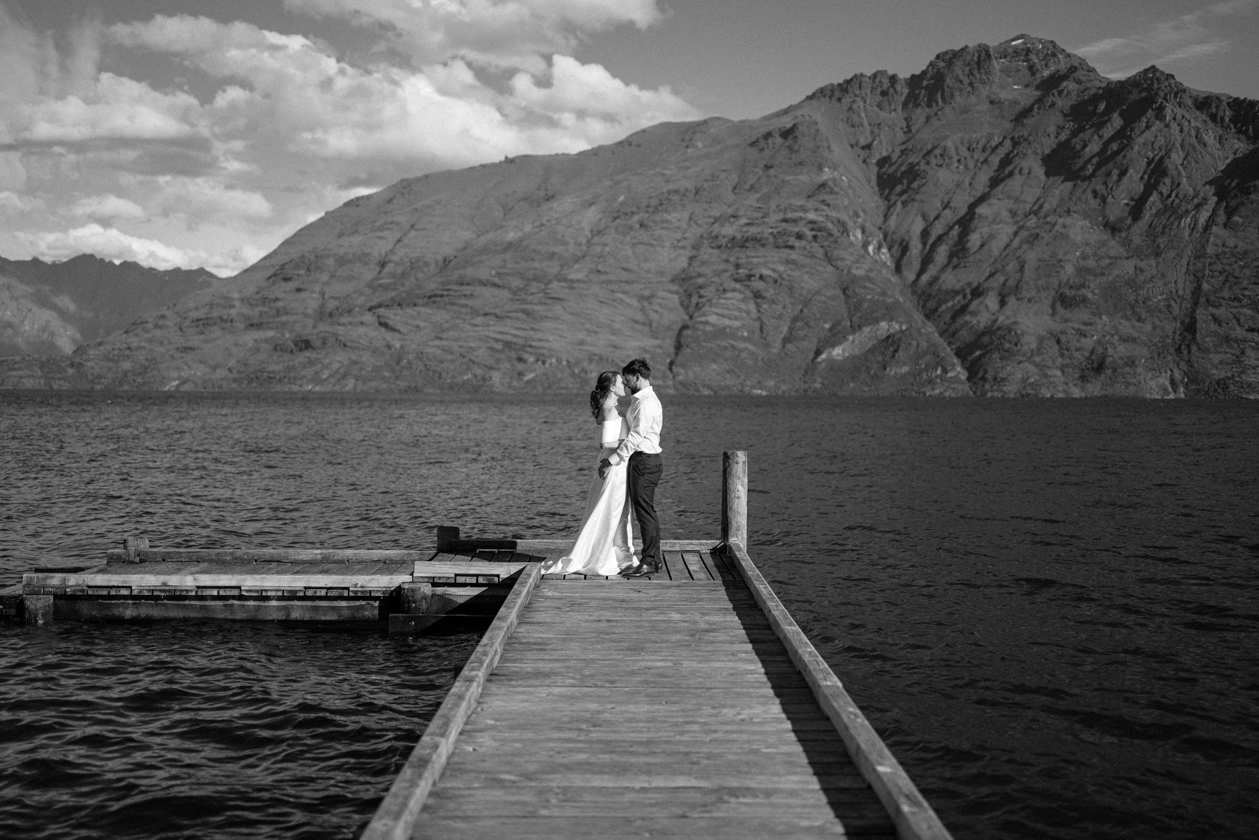 Queenstown Elopement, Otago, New Zealand. A black-and-white photo of a couple in wedding attire standing on a wooden dock at lake Wakatipu, with mountains and clouds in the background.