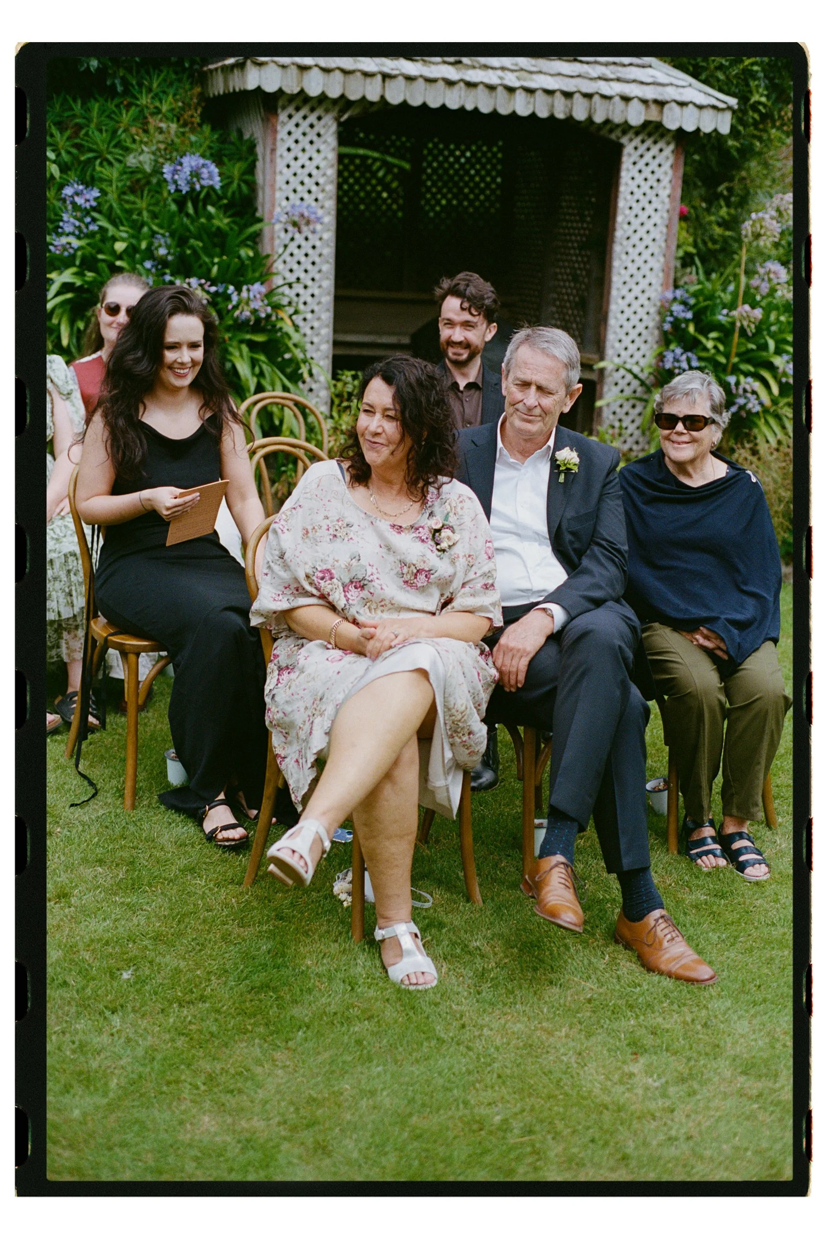 Accrington Farmhouse Wedding, Banks Peninsula, Christchurch, New Zealand. Kodak Portra 400 film photo of wedding guests sitting outdoors on a lawn during a celebration, with a wooden gazebo and green plants with purple flowers in the background.