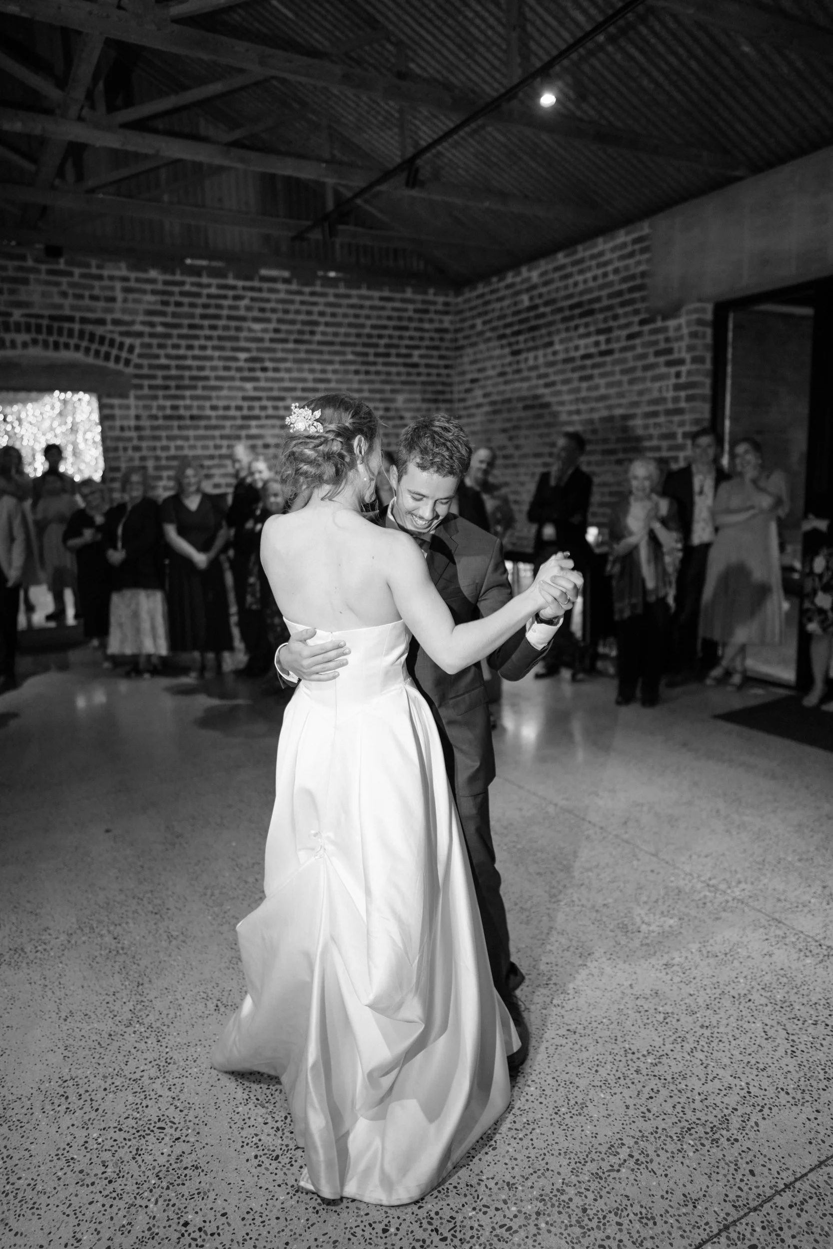 Film + Digital Wedding Photographer Dunedin - Lochend Woolshed, Dunedin, Otago, New Zealand. Bride and groom dancing at their wedding reception. They are smiling and holding each other closely, surrounded by friends and family.