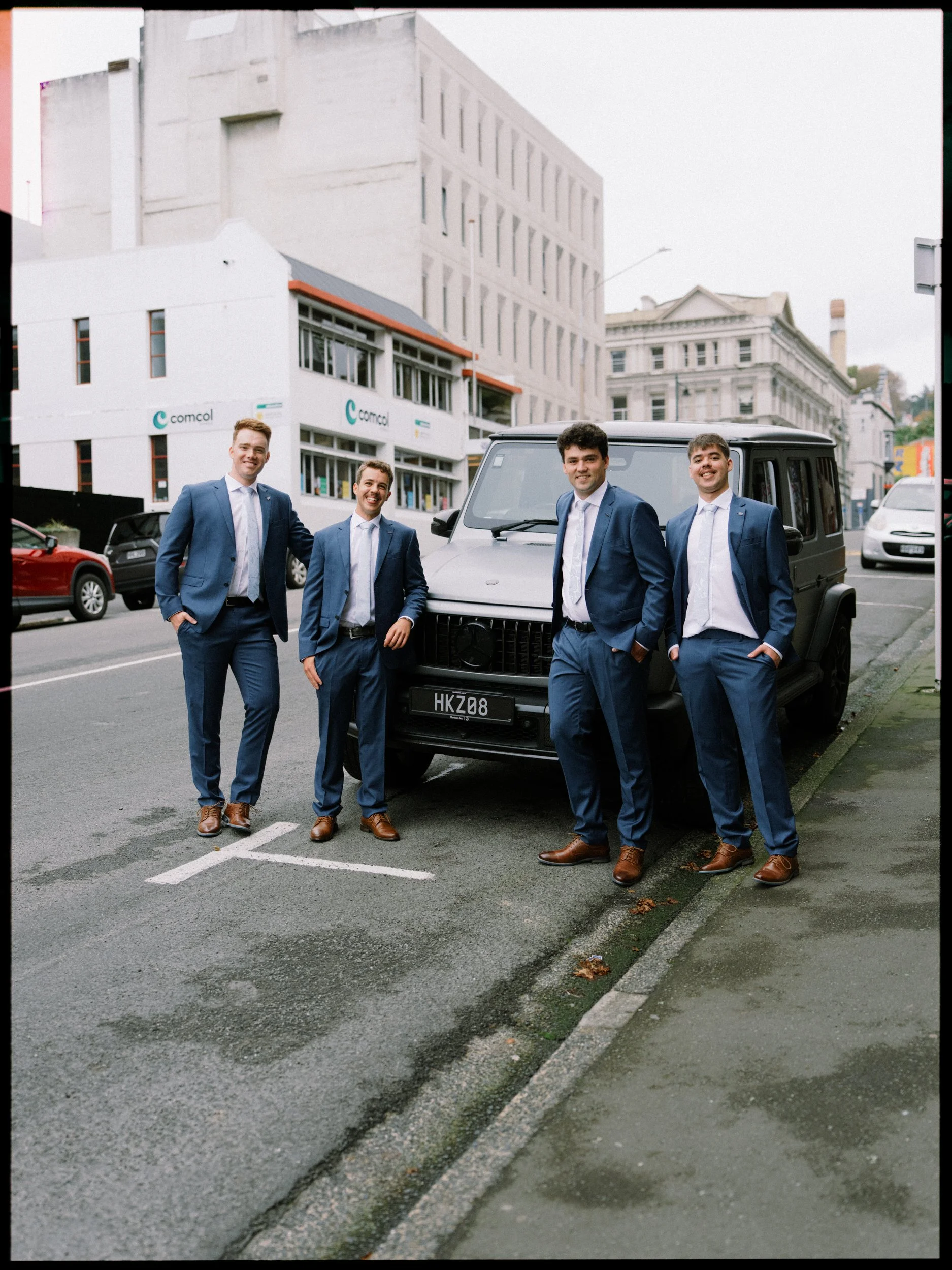 Film + Digital Wedding Photographer Dunedin - Lochend Woolshed, Dunedin, Otago, New Zealand. Groom and groomsmen in blue suits standing on a city street in Dunedin, in front of a Mercedes Benz wagon.
