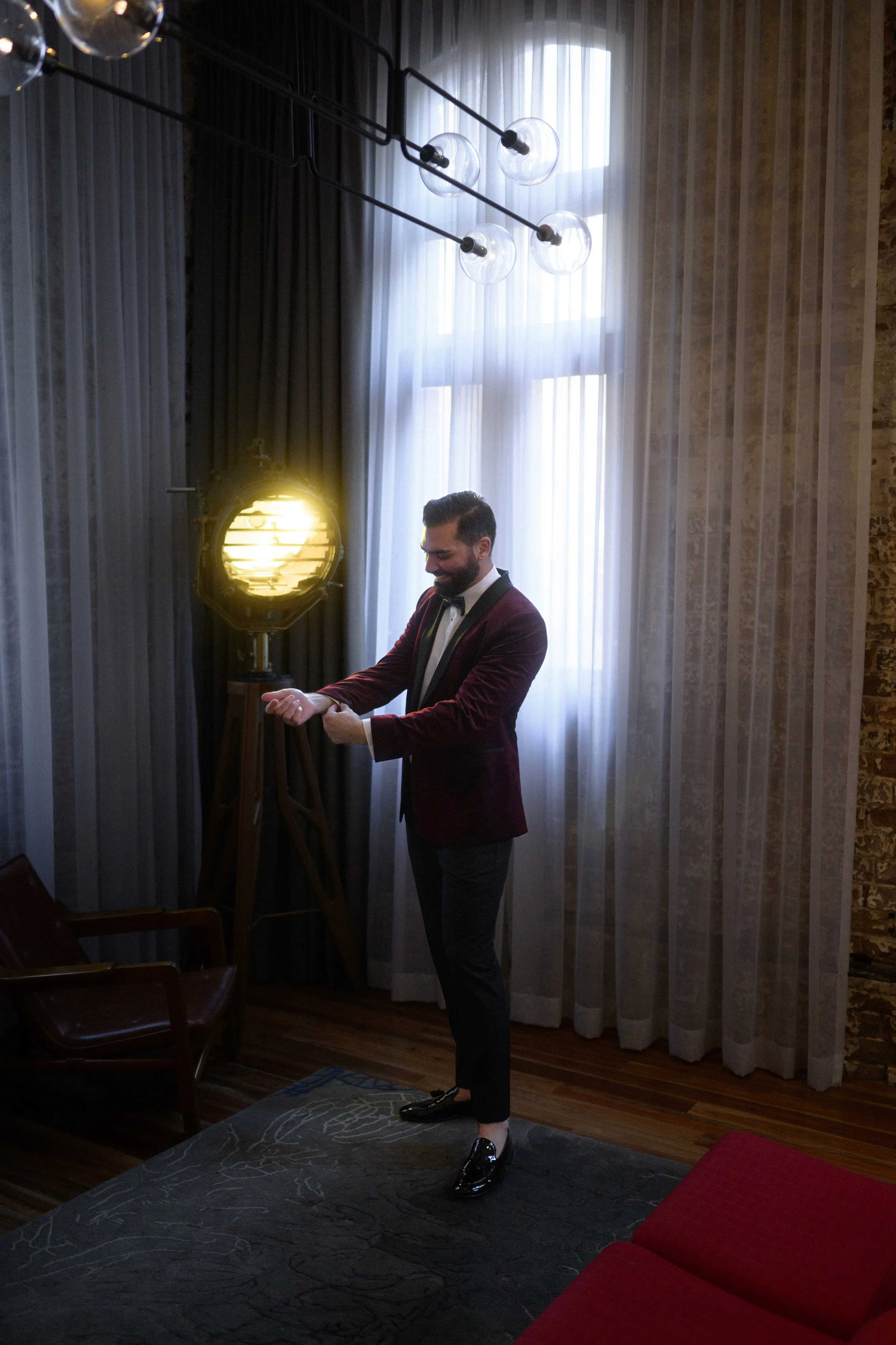 Cinematic wedding photo of groom standing in front of hotel room window and vintage spotlight. He is wearing a burgundy suit and black loafers.
