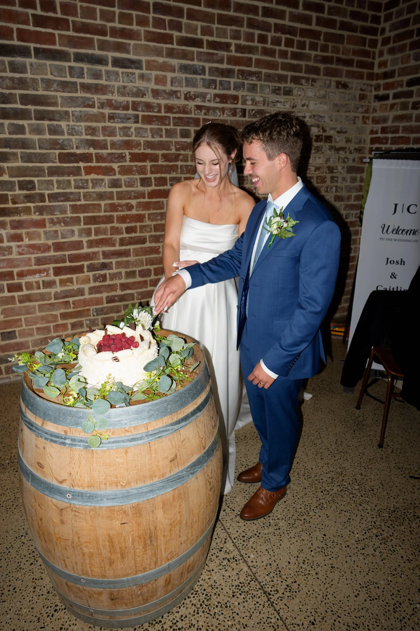 Film + Digital Wedding Photographer Dunedin - Lochend Woolshed, Dunedin, Otago, New Zealand. Bride and groom smiling and cutting wedding cake with white flowers and raspberries on a wine barrel in a rustic setting with a brick wall.