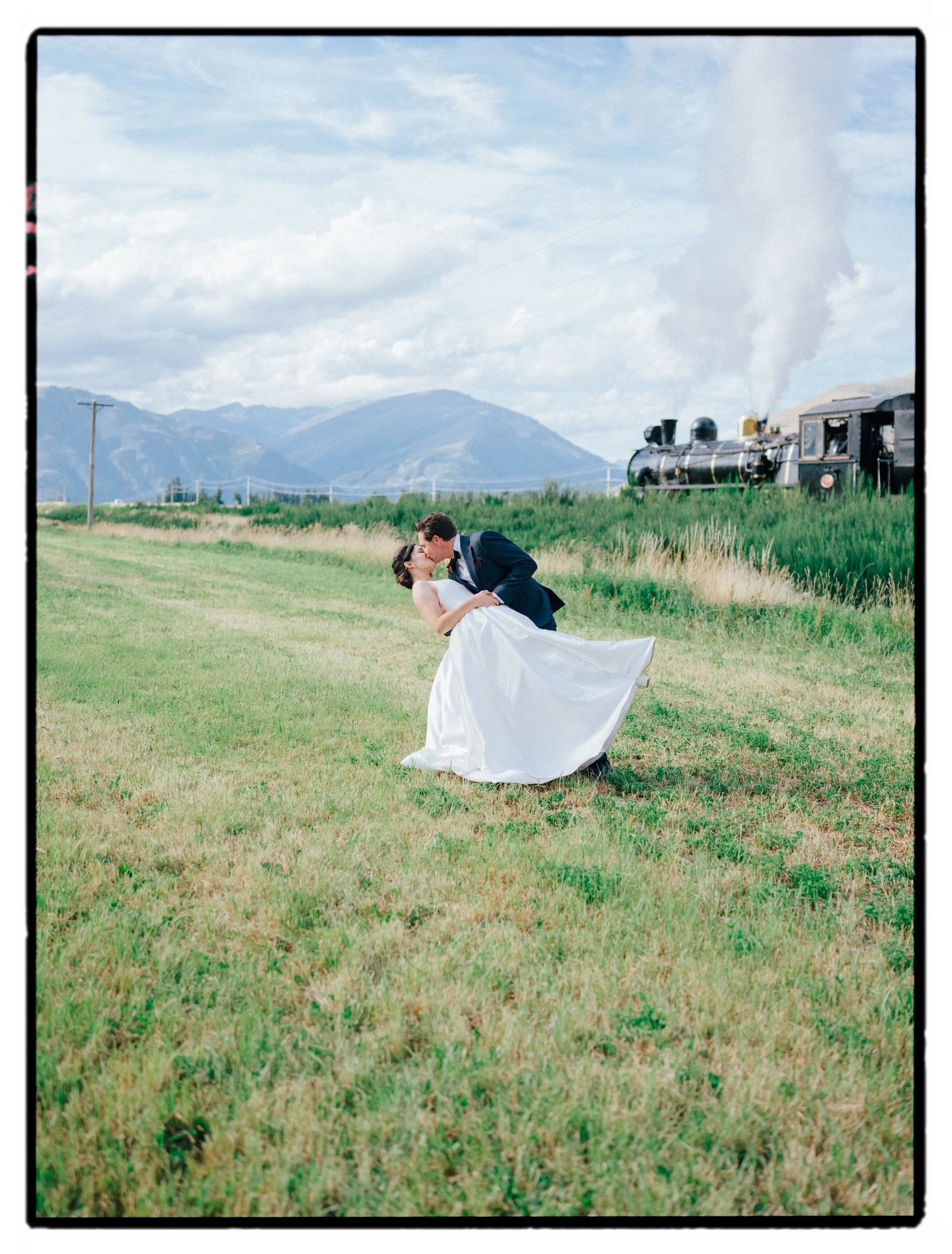 Film + Digital Wedding Photographer Christchurch - Kingston Wedding, Otago, New Zealand. Cinematic photo of bride and groom kissing on a grass field with mountains and Kingston Flyer steam train in the background.