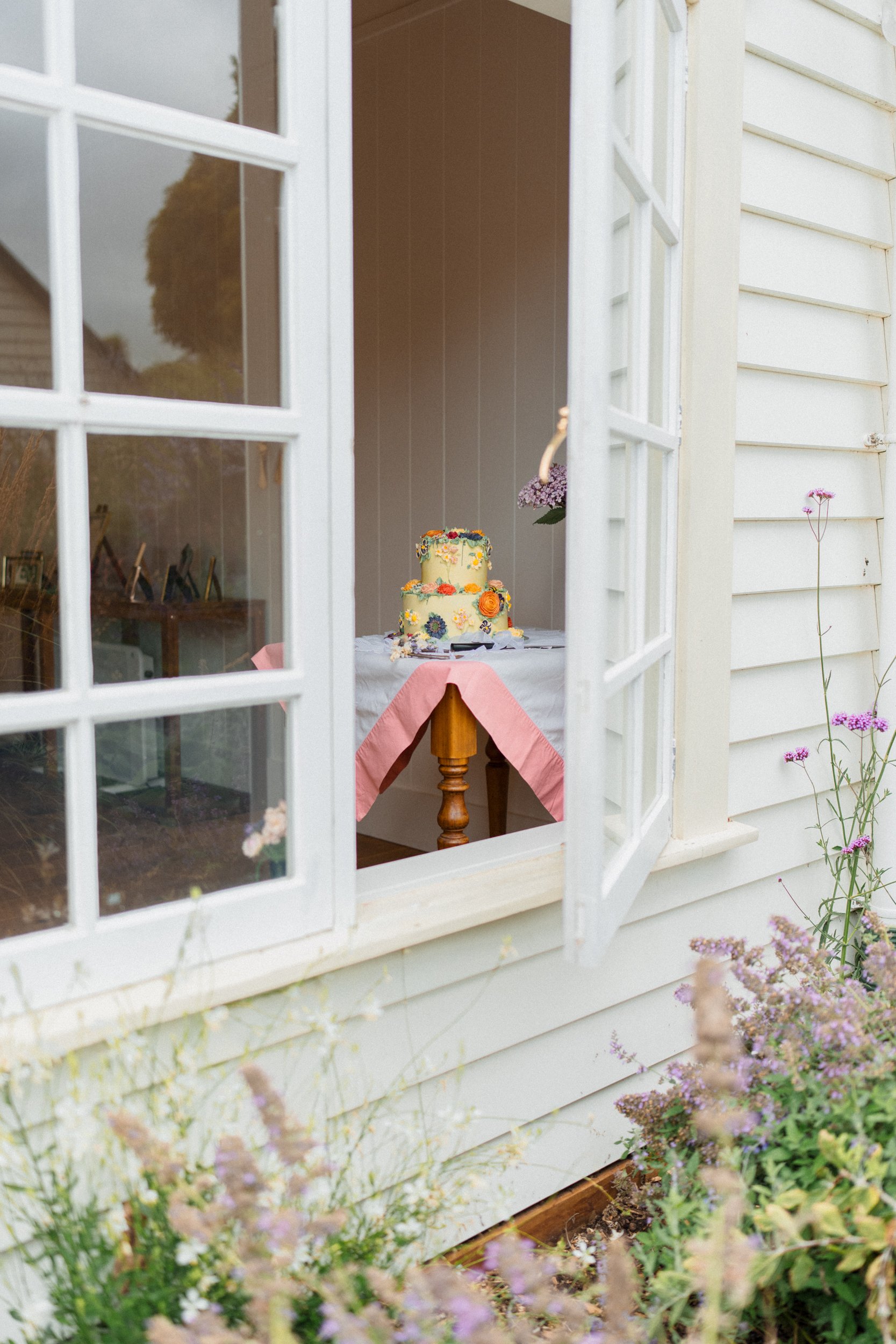 Accrington Farmhouse Wedding, Banks Peninsula, Christchurch, New Zealand. Yellow wedding cake on a table, decorated with flowers, seen through an open window of a barn with white siding and purple flowers outside.