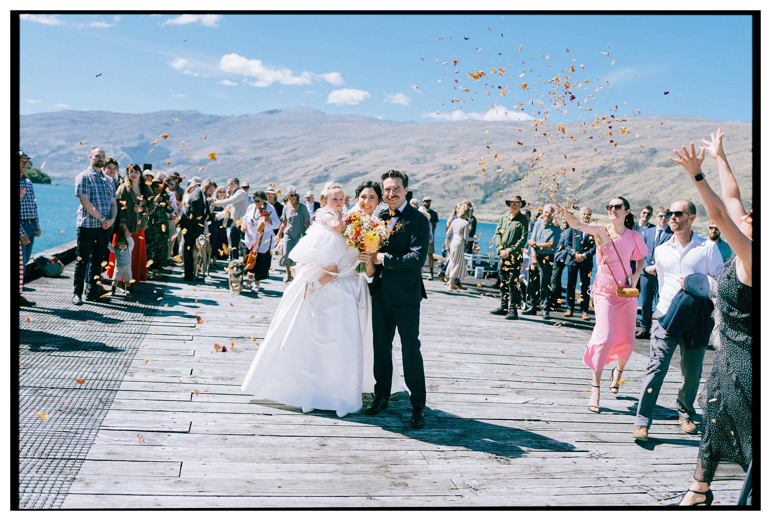 Kingston Wedding, Otago, New Zealand. Film photo of bride and groom with their baby daughter smiling after ceremony at the lake wharf surrounded by guests throwing flower petals at them and mountains in the background.