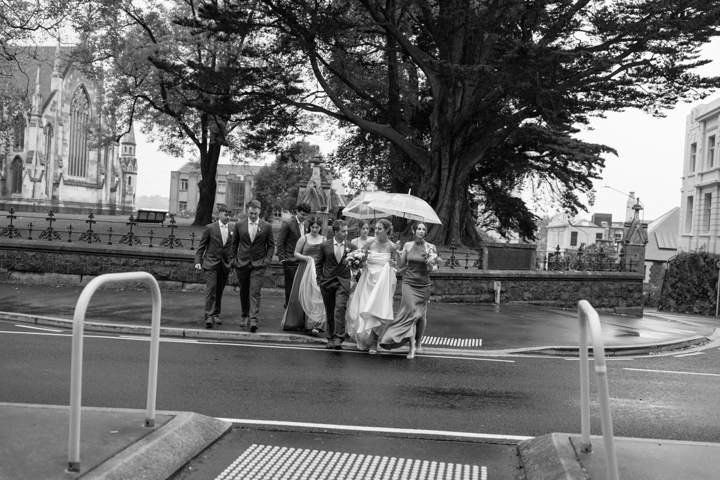 Film + Digital Wedding Photographer Dunedin - Lochend Woolshed, Dunedin, Otago, New Zealand. Black and white photo of bridal party crossing a street, holding umbrellas, with a large tree and historic buildings in the background.