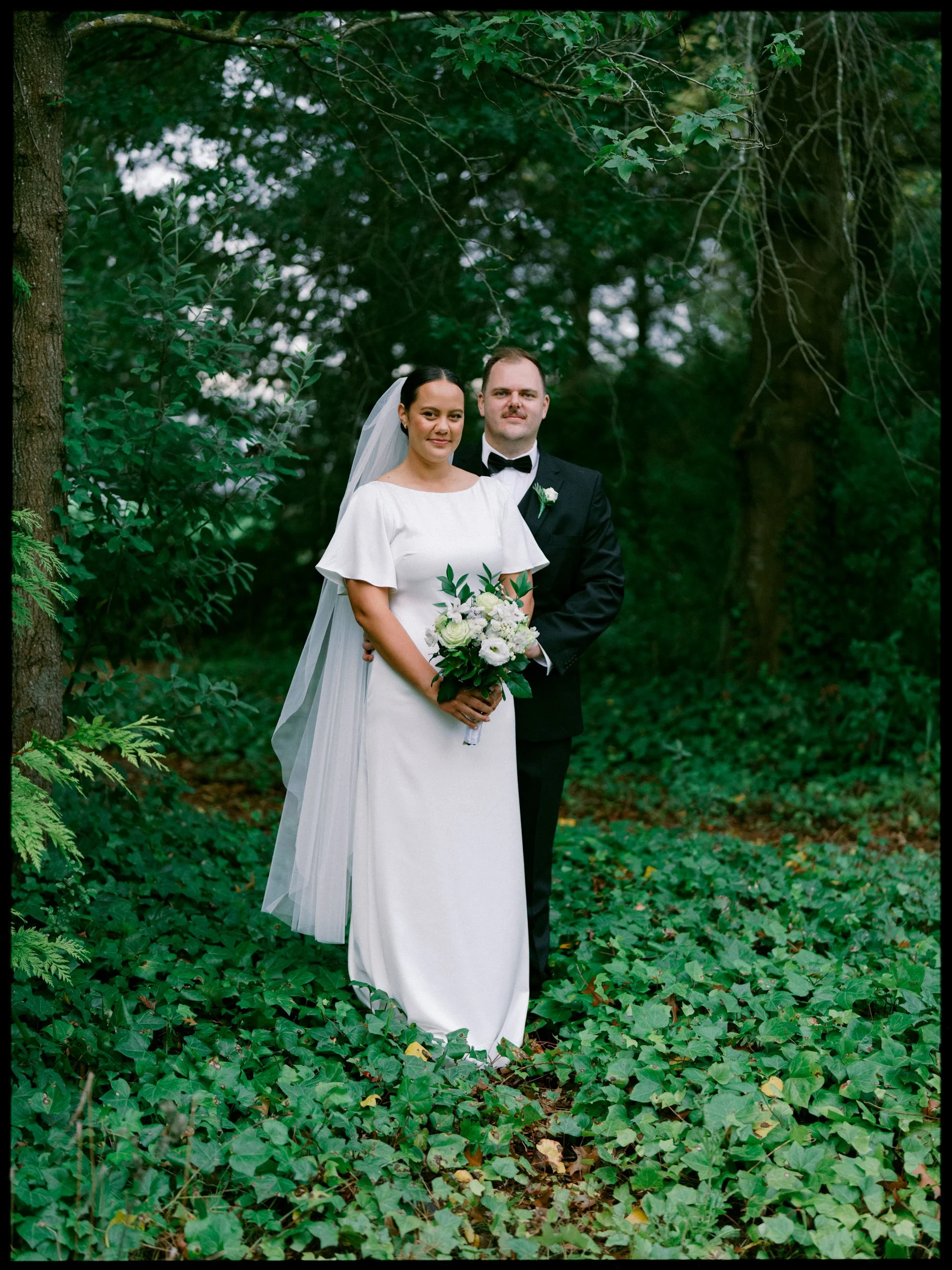 Film + Digital Wedding Photographer Christchurch - Backyard Wedding, Christchurch, Canterbury, New Zealand. Bride and groom standing outdoors in a lush, green, woodland setting. The background is filled with trees and dense foliage.