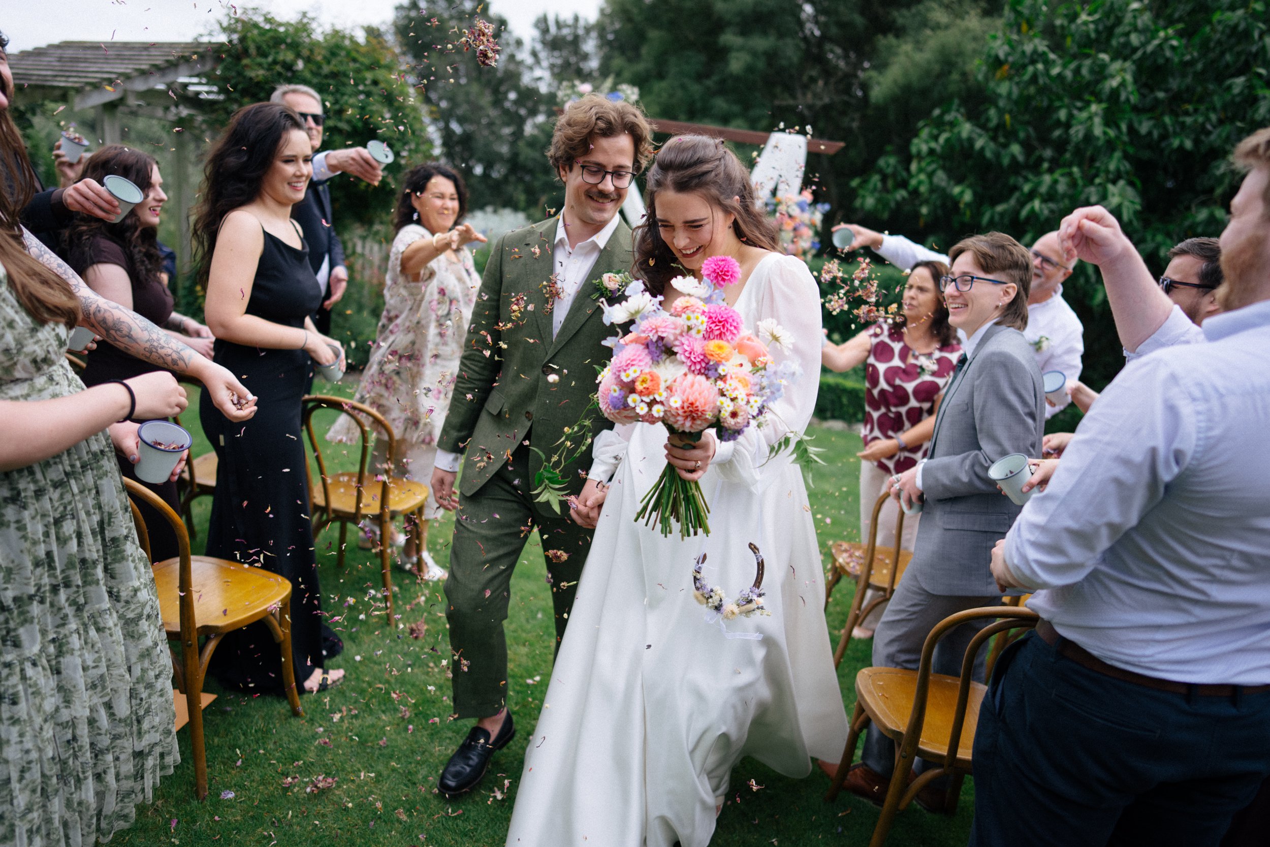 Accrington Farmhouse Wedding, Banks Peninsula, Christchurch, New Zealand. Bride holding a bouquet of pink and white flowers, walking hand-in-hand with groom, surrounded by guests throwing flower petals at an outdoor wedding ceremony.