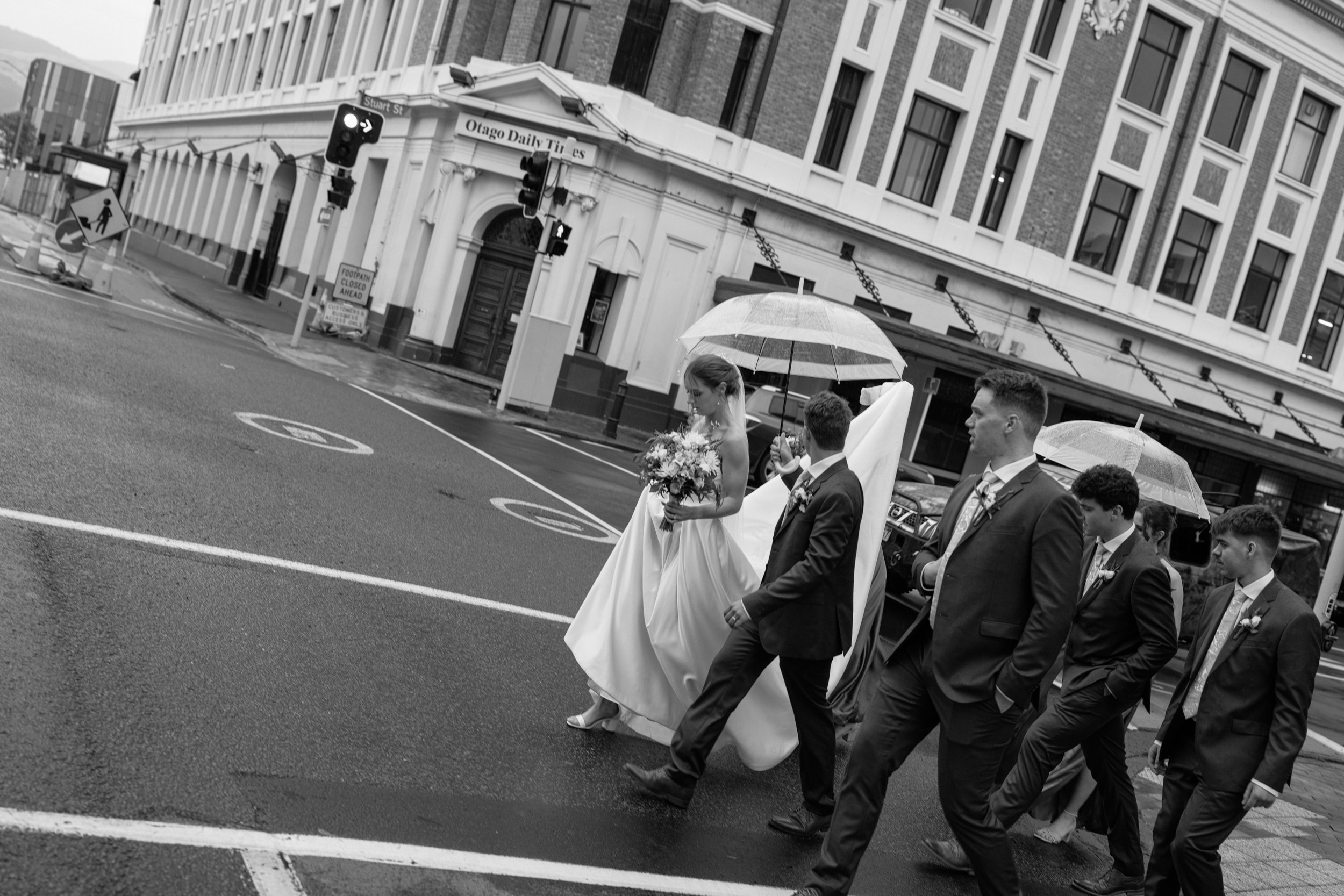 Film + Digital Wedding Photographer Dunedin - Lochend Woolshed, Dunedin, Otago, New Zealand. Bridal party walk across city street on rainy day, holding umbrellas and bride holding a bouquet, against the backdrop of tall buildings and traffic signals.