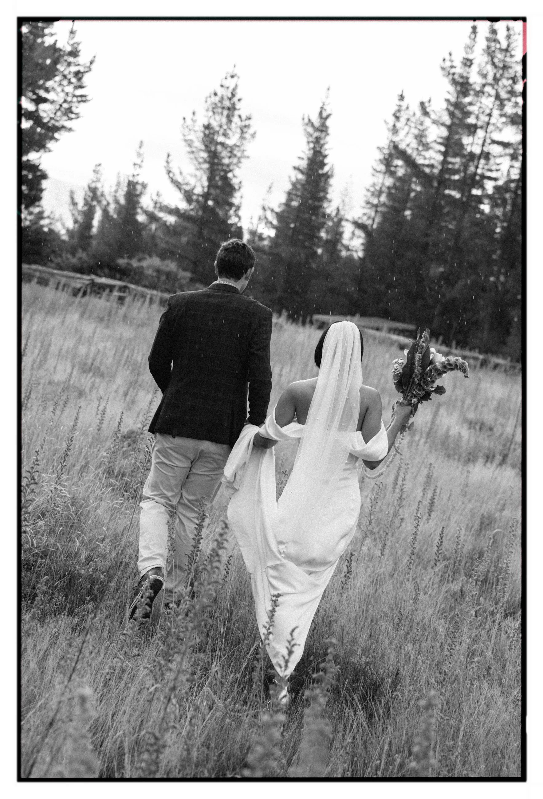Film + Digital Wedding Photographer Christchurch - Alexandra Wedding, Otago, New Zealand. Black and white photo of bride and groom walking in pine forest on a rainy day. Bride is holding her wedding dress up and a bouquet of flowers.
