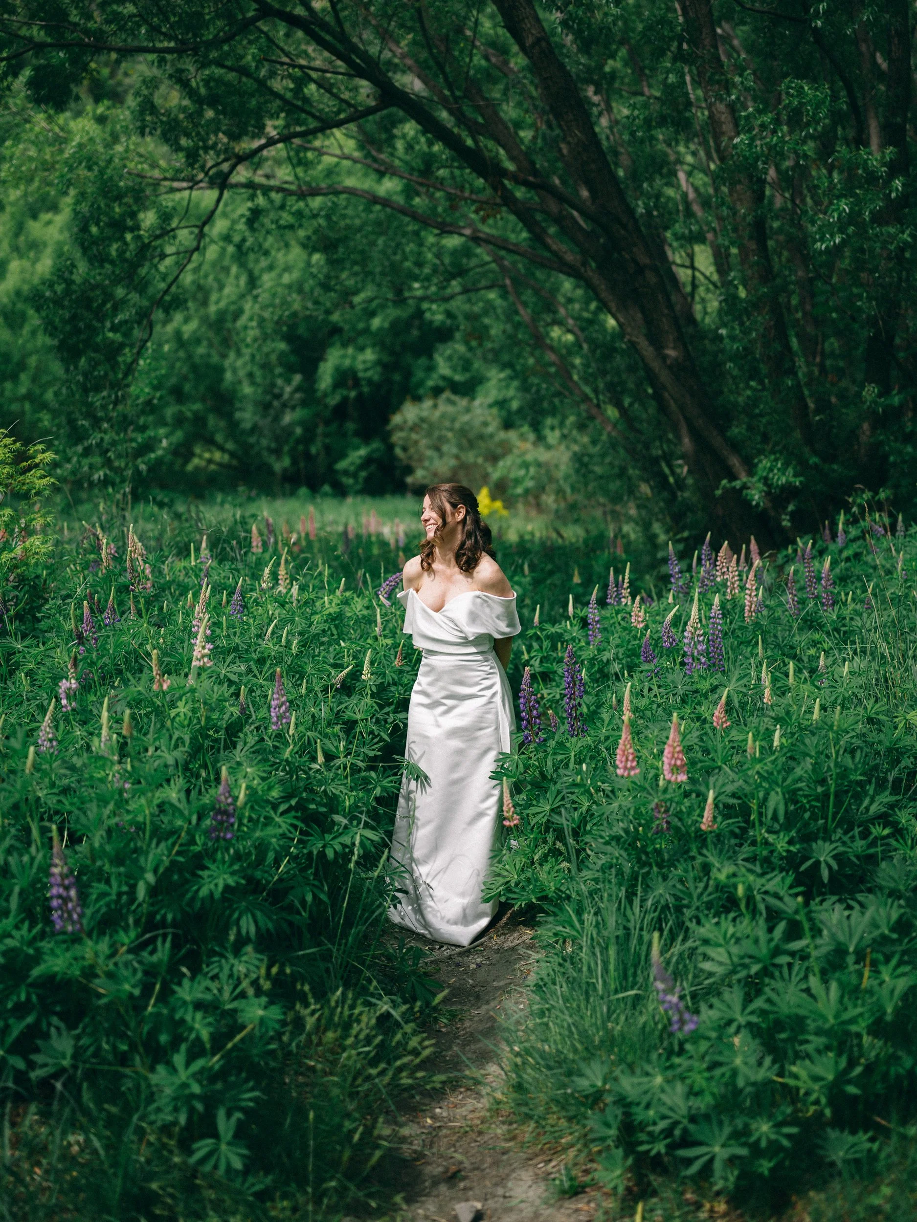 Queenstown Elopement, Otago, New Zealand. Bride in white wedding dress stands on a path near the Arrow river in Arrowtown in a lush green forest surrounded by tall lupine flowers in purple, pink, and cream colors, with large trees overhead.
