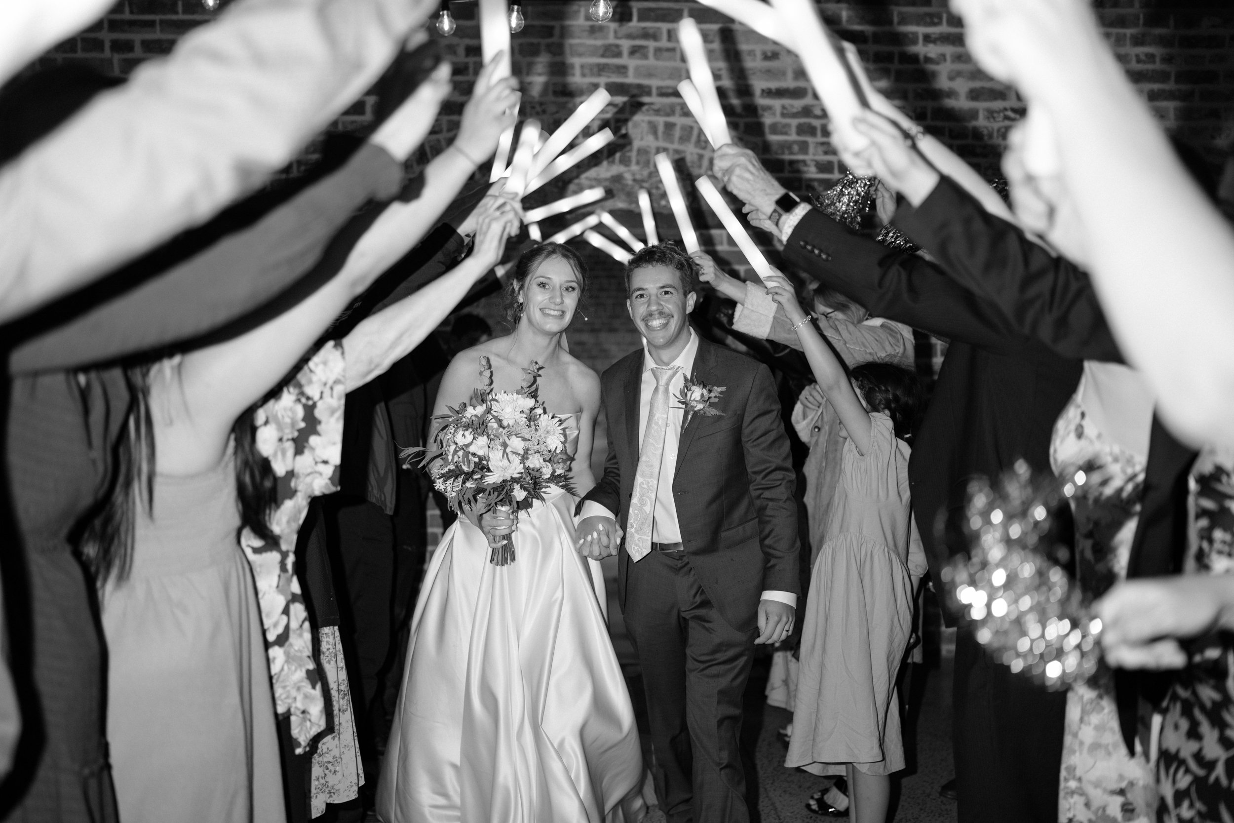Film + Digital Wedding Photographer Dunedin - Lochend Woolshed, Dunedin, Otago, New Zealand. Black and white photo of bride and groom walking hand in hand through a tunnel of people's arms holding light sticks entering wedding reception.