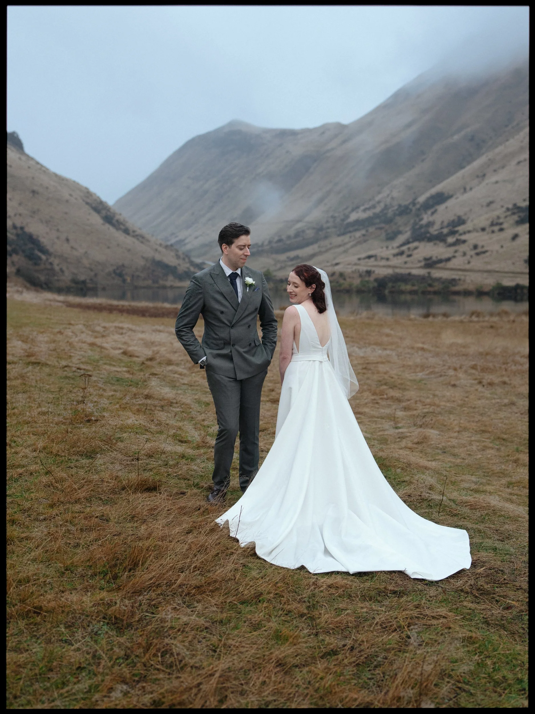 Film + Digital Wedding Photographer Queenstown - Kāmana Lakehouse, Queenstown, Otago, New Zealand. Bride and groom standing on a grassy field with mountains in the background at Moke Lake. Moody rainy wedding photography.
