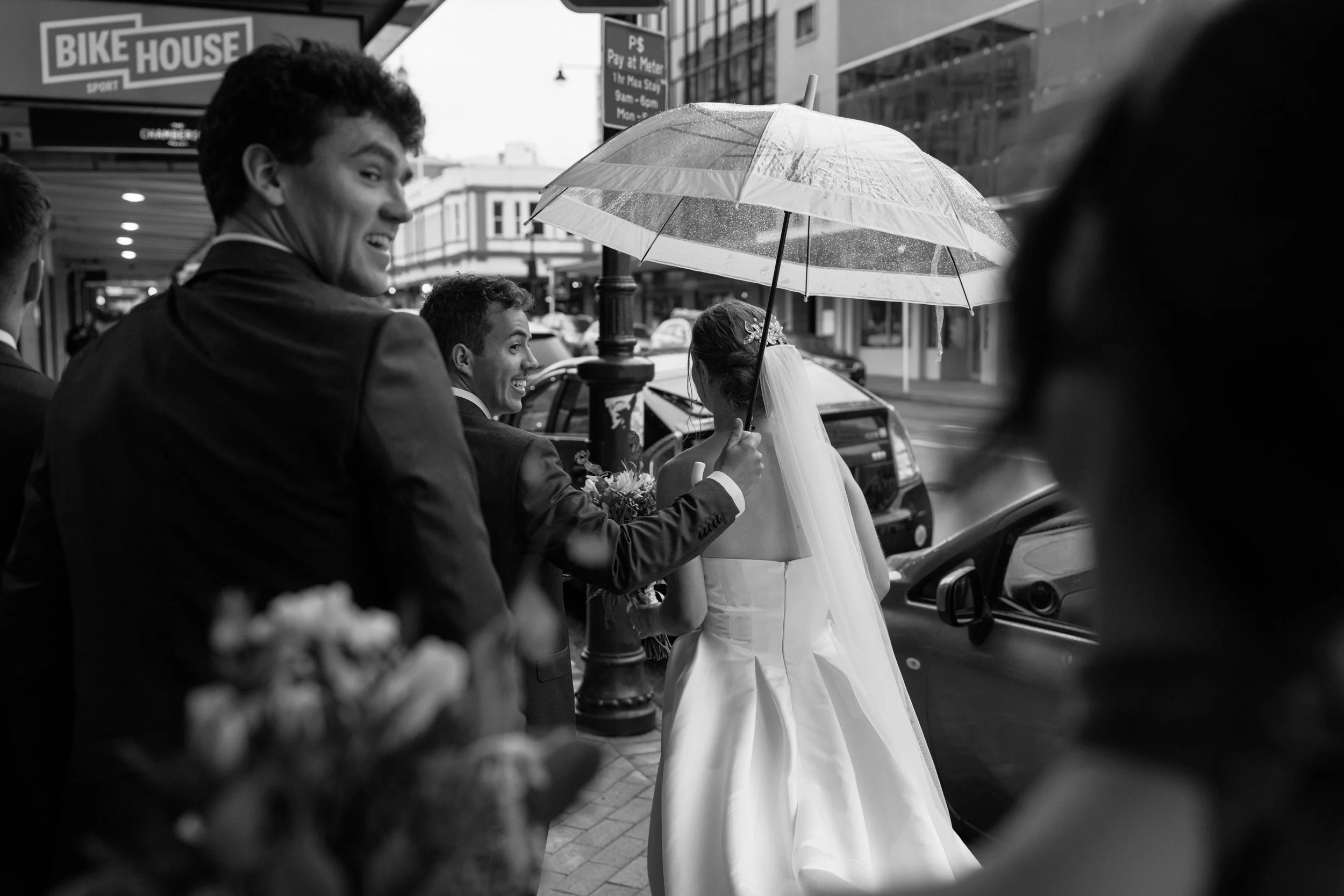 Film + Digital Wedding Photographer Dunedin - Lochend Woolshed, Dunedin, Otago, New Zealand. Black and white photo of a bride and groom standing on a city street, with the groom holding umbrella over bride while they smile and look at each other.