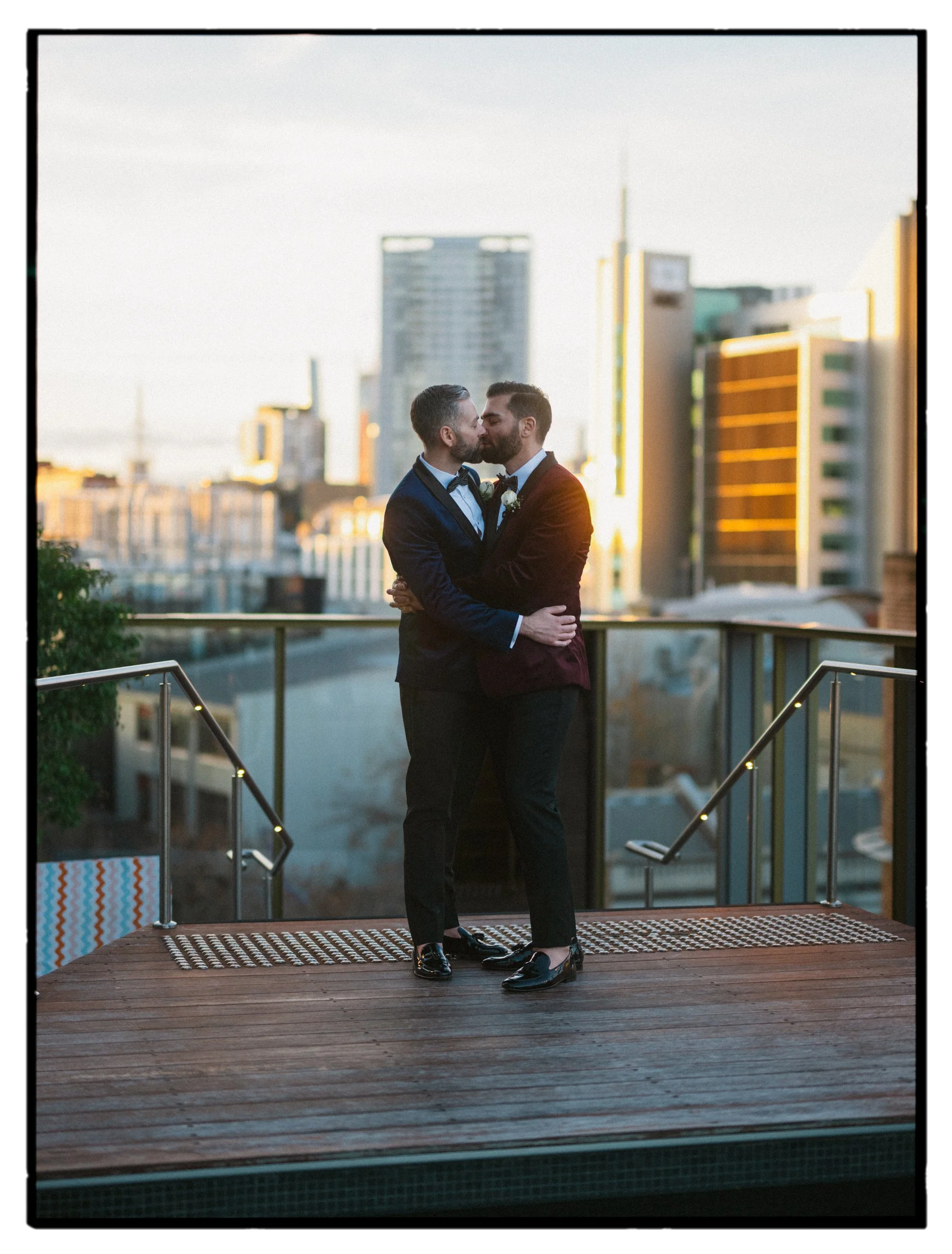 Christchurch, Queenstown, Dunedin Film + Digital wedding photography. Sunset photo of grooms on terrace kissing with city skyline in the background