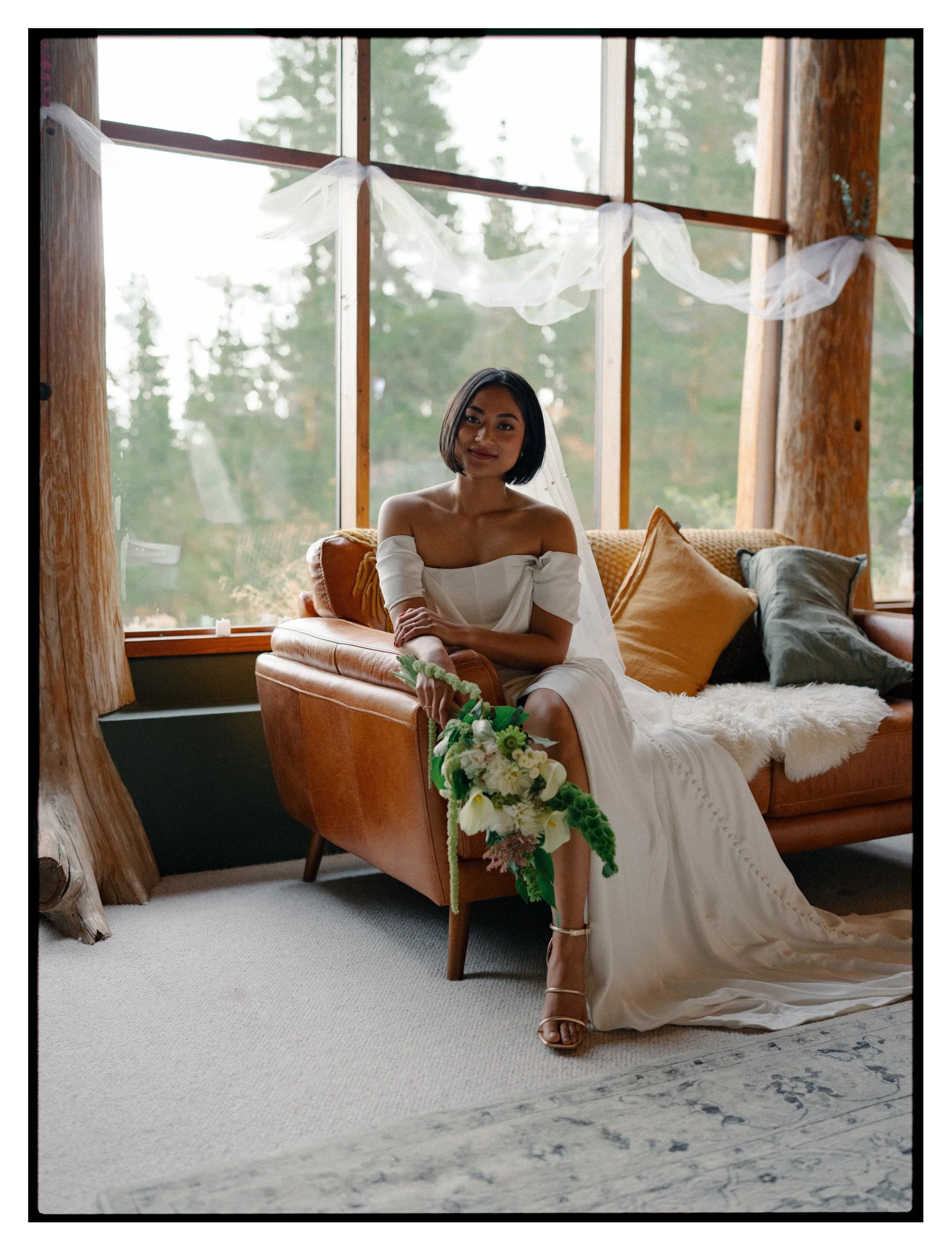 Film + Digital Wedding Photographer Christchurch - Alexandra Wedding, Otago, New Zealand. Film photo of bride sitting on leather lounge holding a bouquet of white flowers inside Fairytale Log Cabin at Homewood Forest Retreat.
